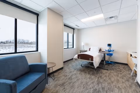Bright interior hospital patient room with a bed, blue couch, windows, and equipment.