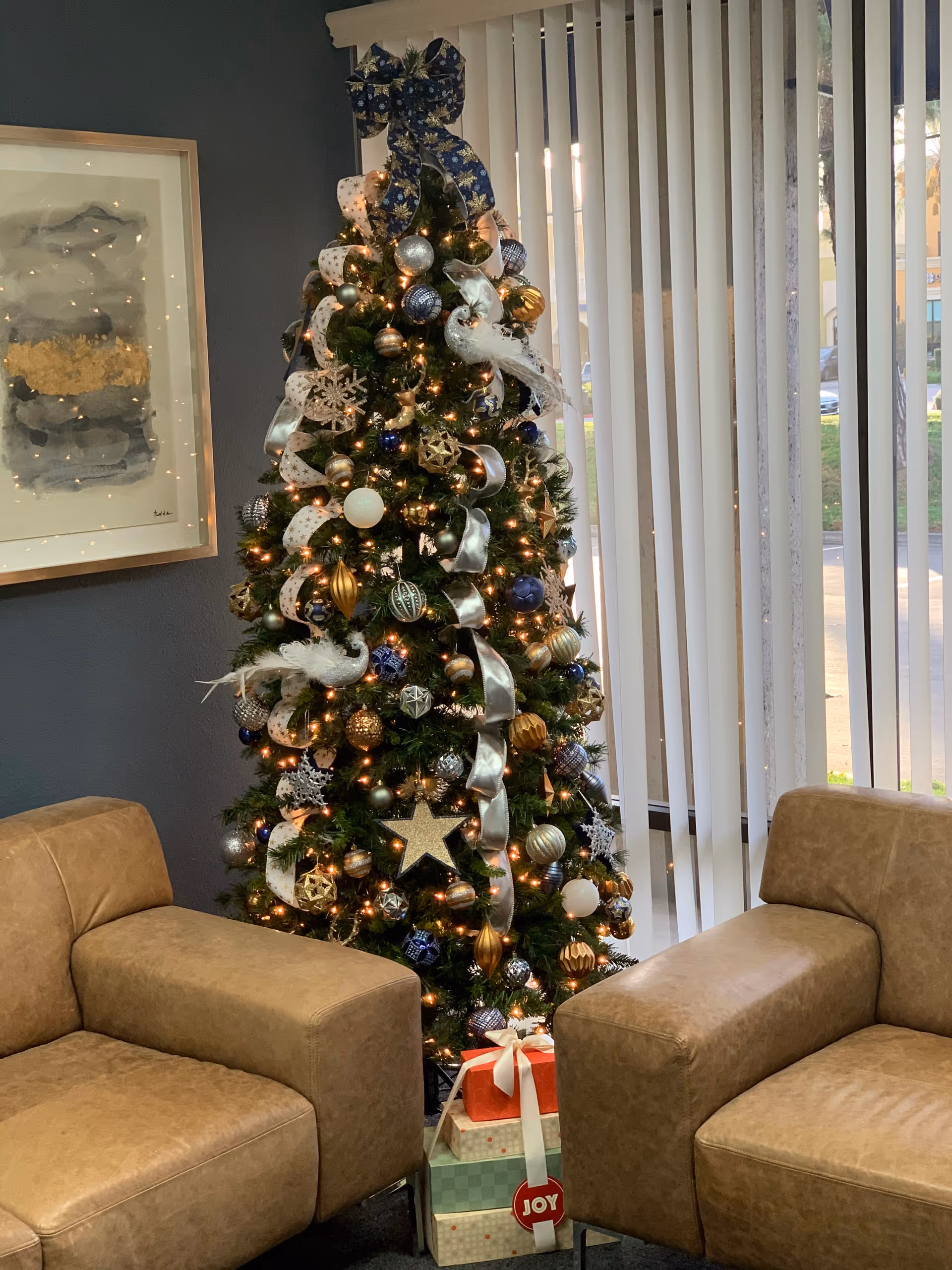 A decorated Christmas tree stands beside beige lounge chairs in a facility lobby.