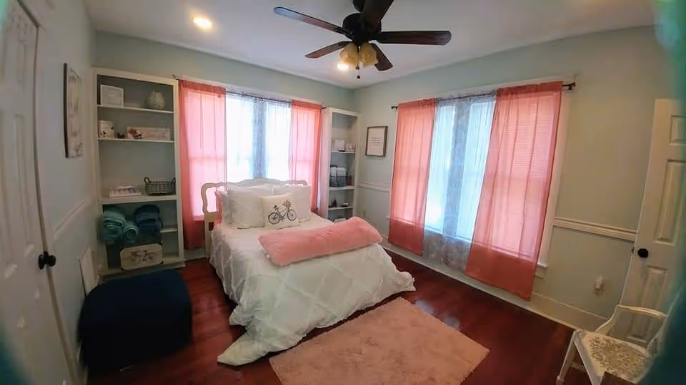 Cozy light-blue bedroom with white bed, pink curtains, shelves, and pink rug.