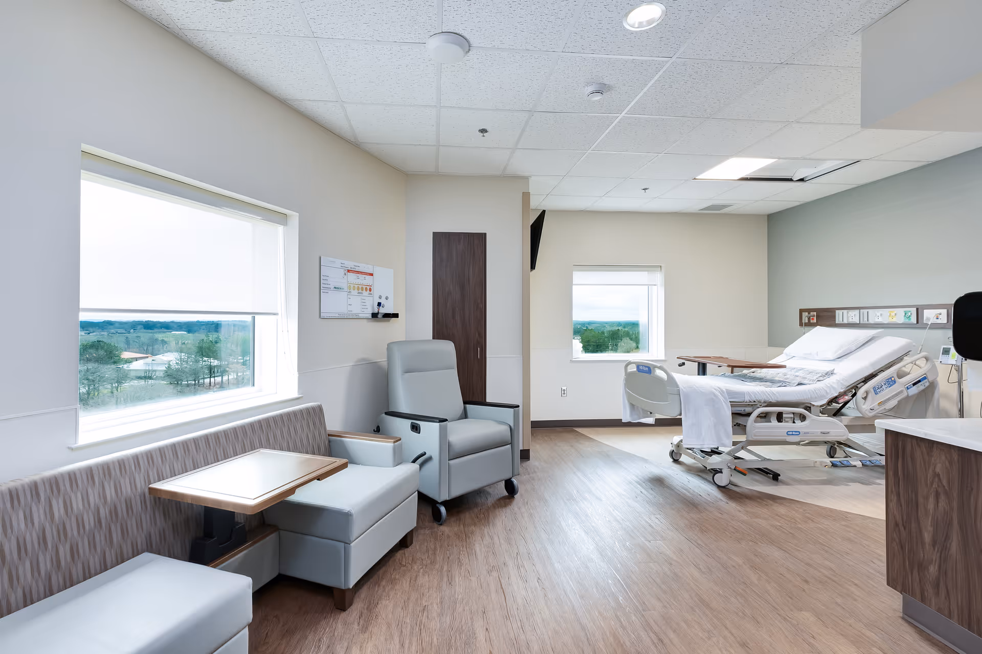 Bright hospital room featuring a patient bed, seating area, and large windows.