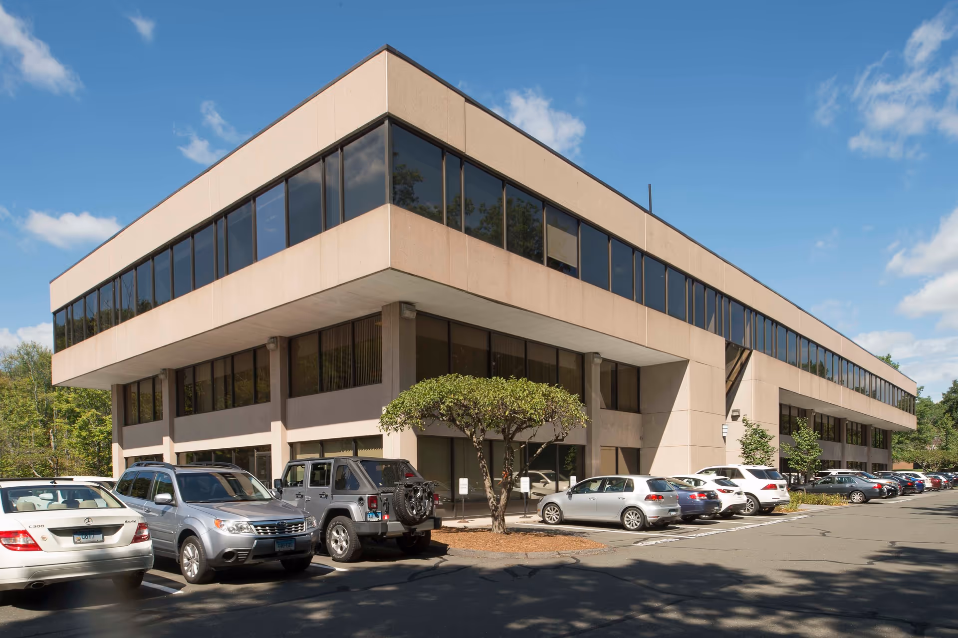 Two-story beige building with large glass windows and a parking lot in front.