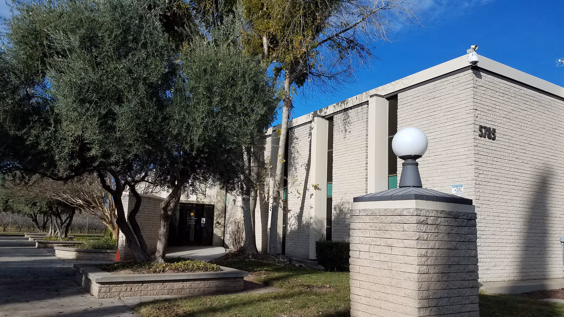 Exterior view of a beige brick facility entrance with trees and blue sky.