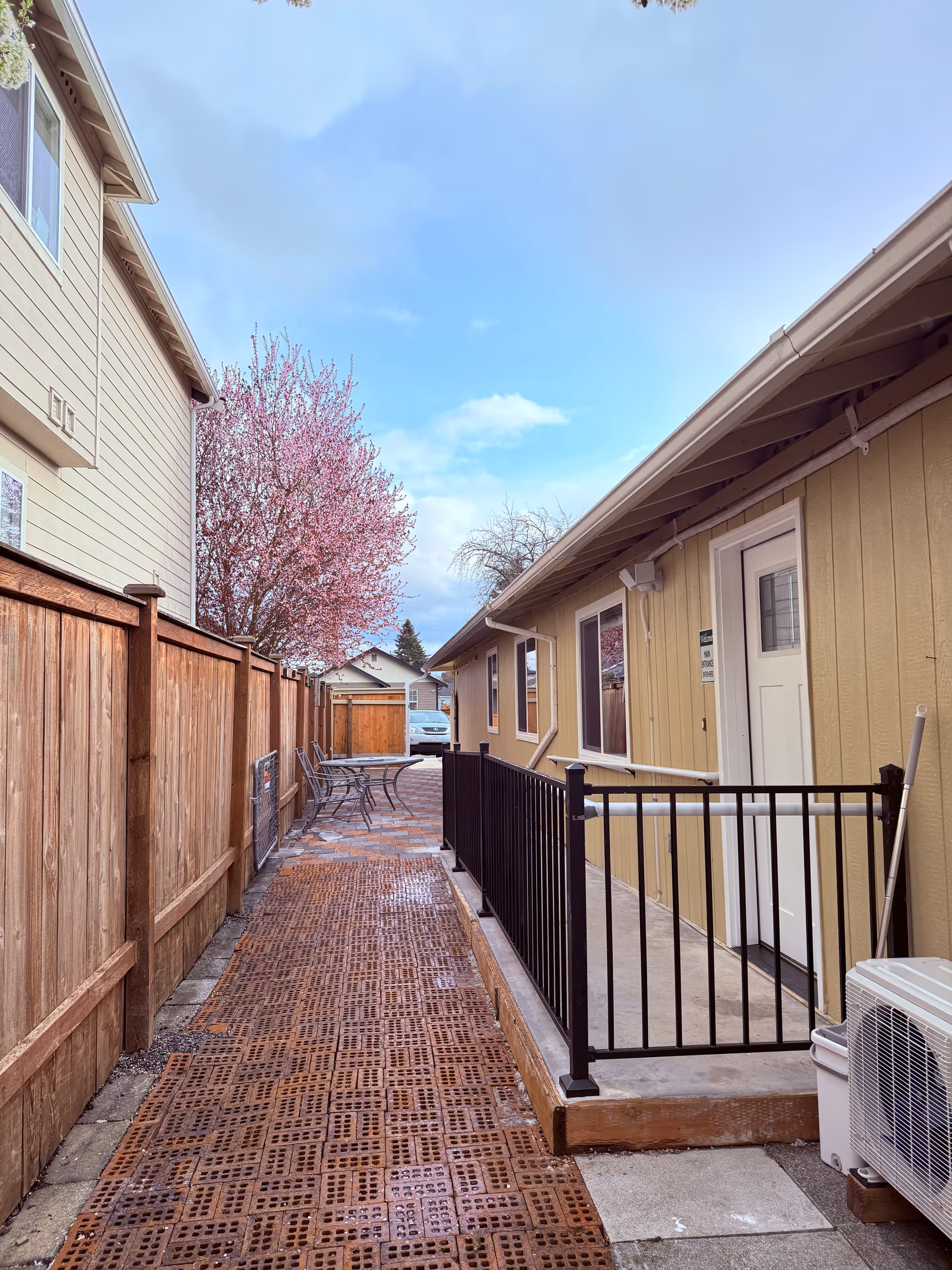 Outdoor walkway between beige buildings, pink blossom tree, railing, table and chairs along the path.