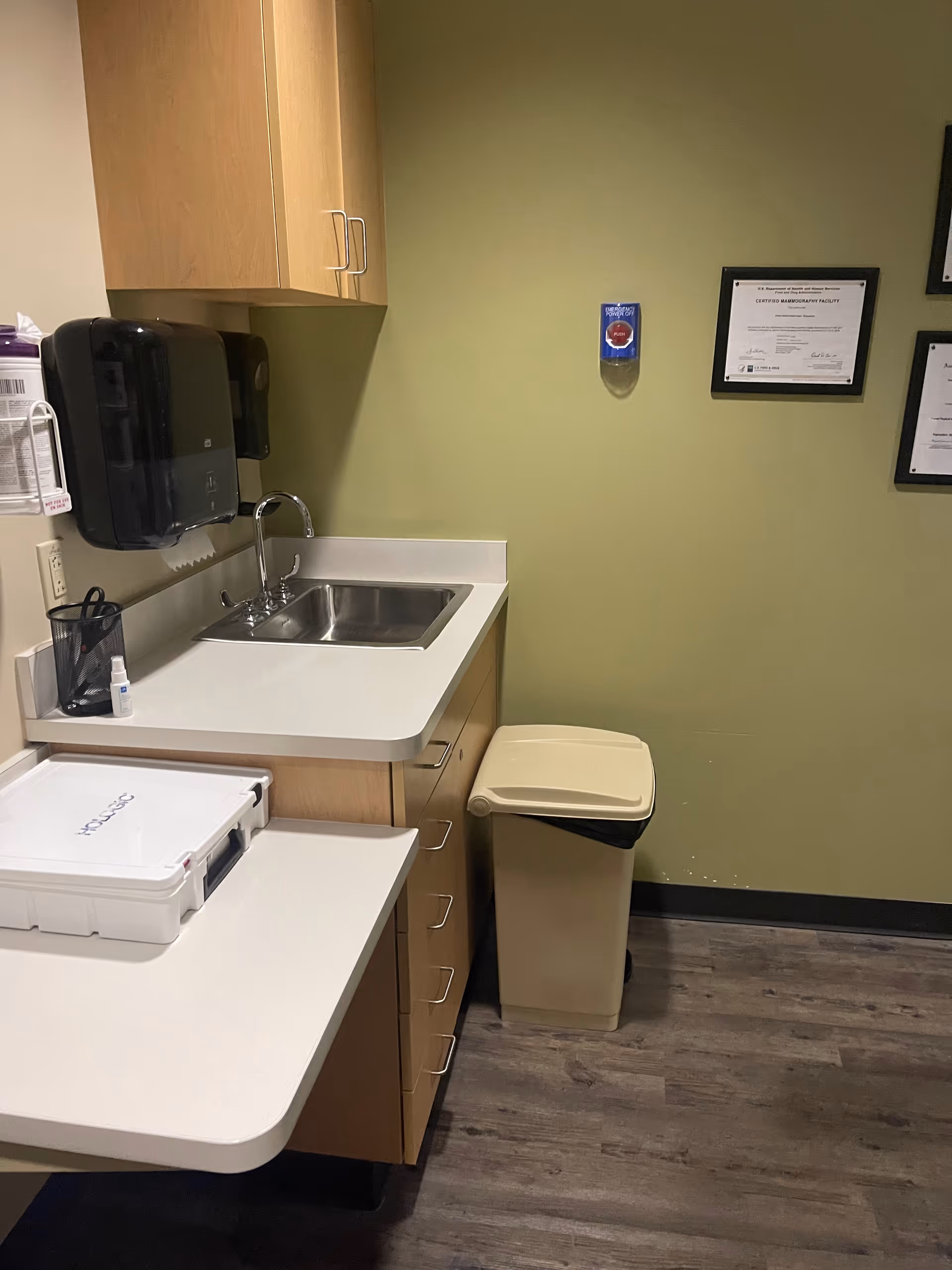 Small medical facility kitchenette with sink, cabinets, trash can, and framed certificates on green wall.