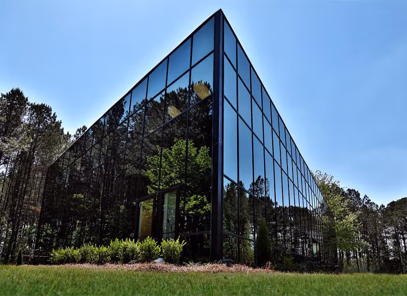 Modern glass-front building with reflective windows surrounded by trees and grass.