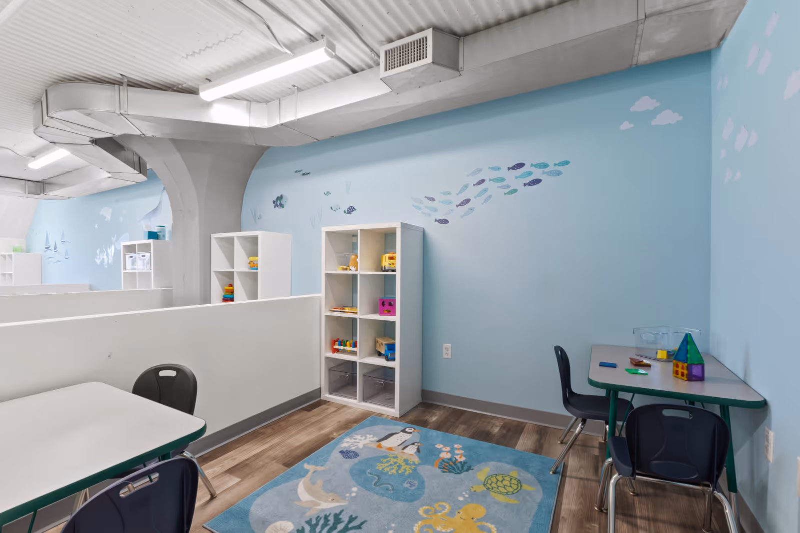 Indoor play area with blue walls, shelves of toys, and small tables.