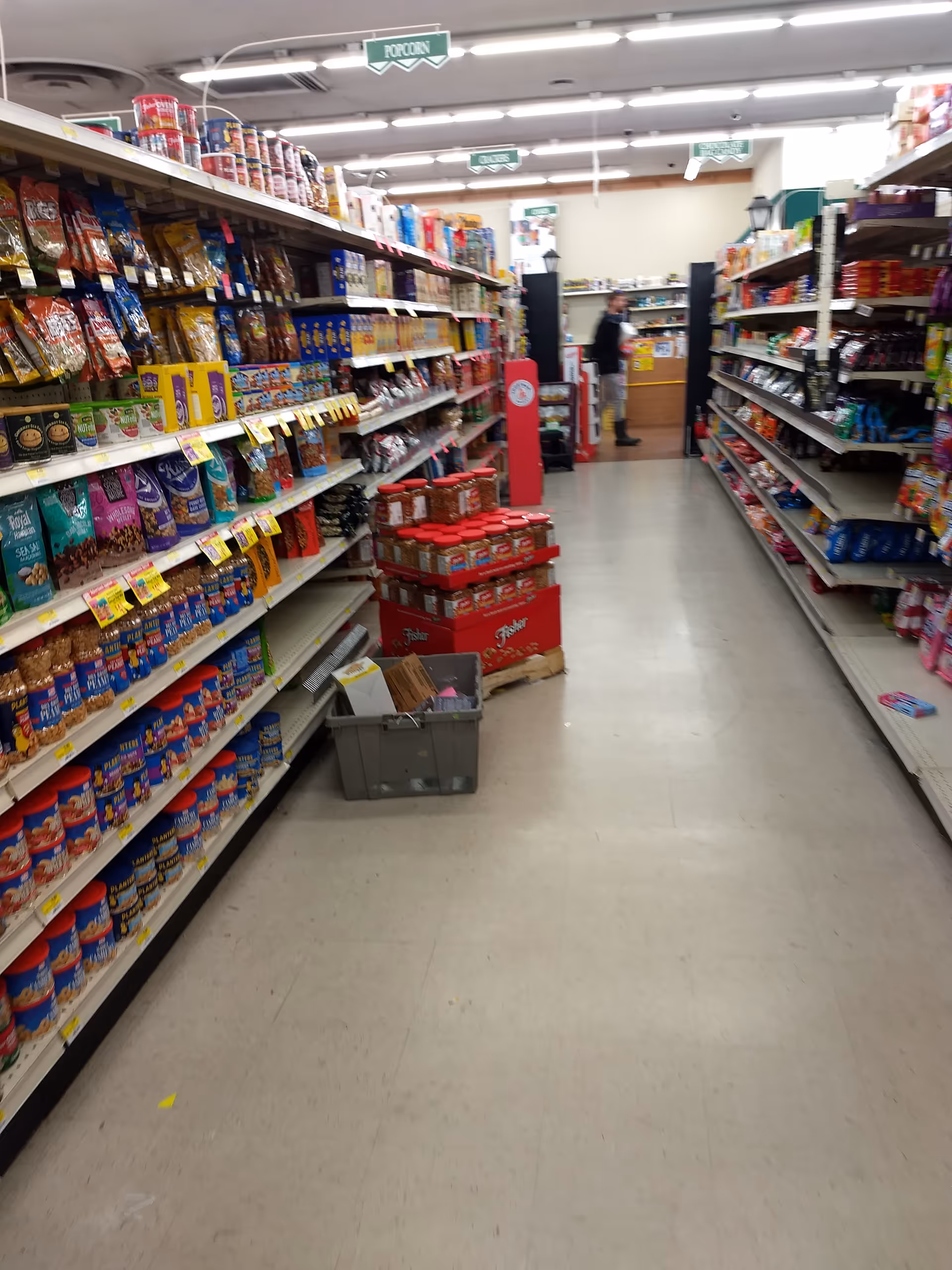 A grocery store aisle filled with snack shelves and a shopper in the background.