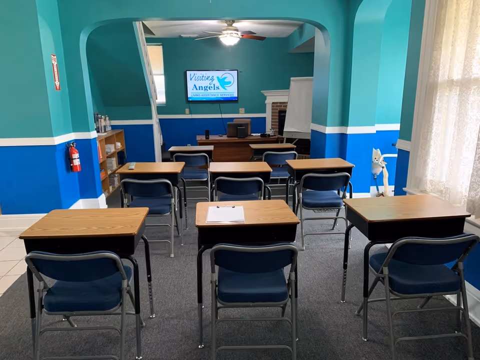 Classroom-style training room with desks, chairs, and a Visiting Angels screen at the front