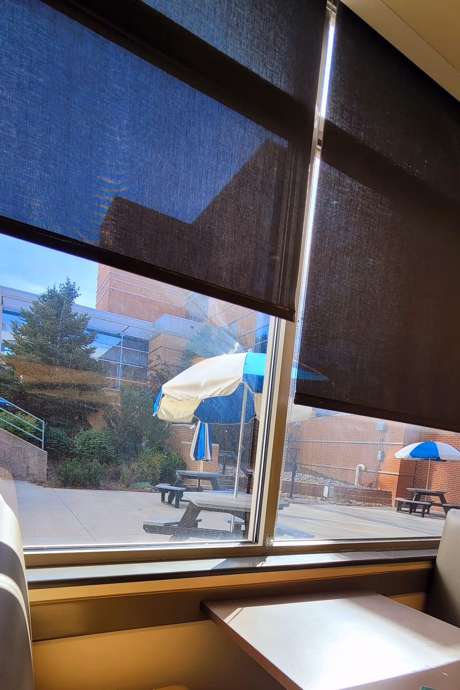 Indoor window view of an outdoor courtyard with picnic tables and blue umbrellas.