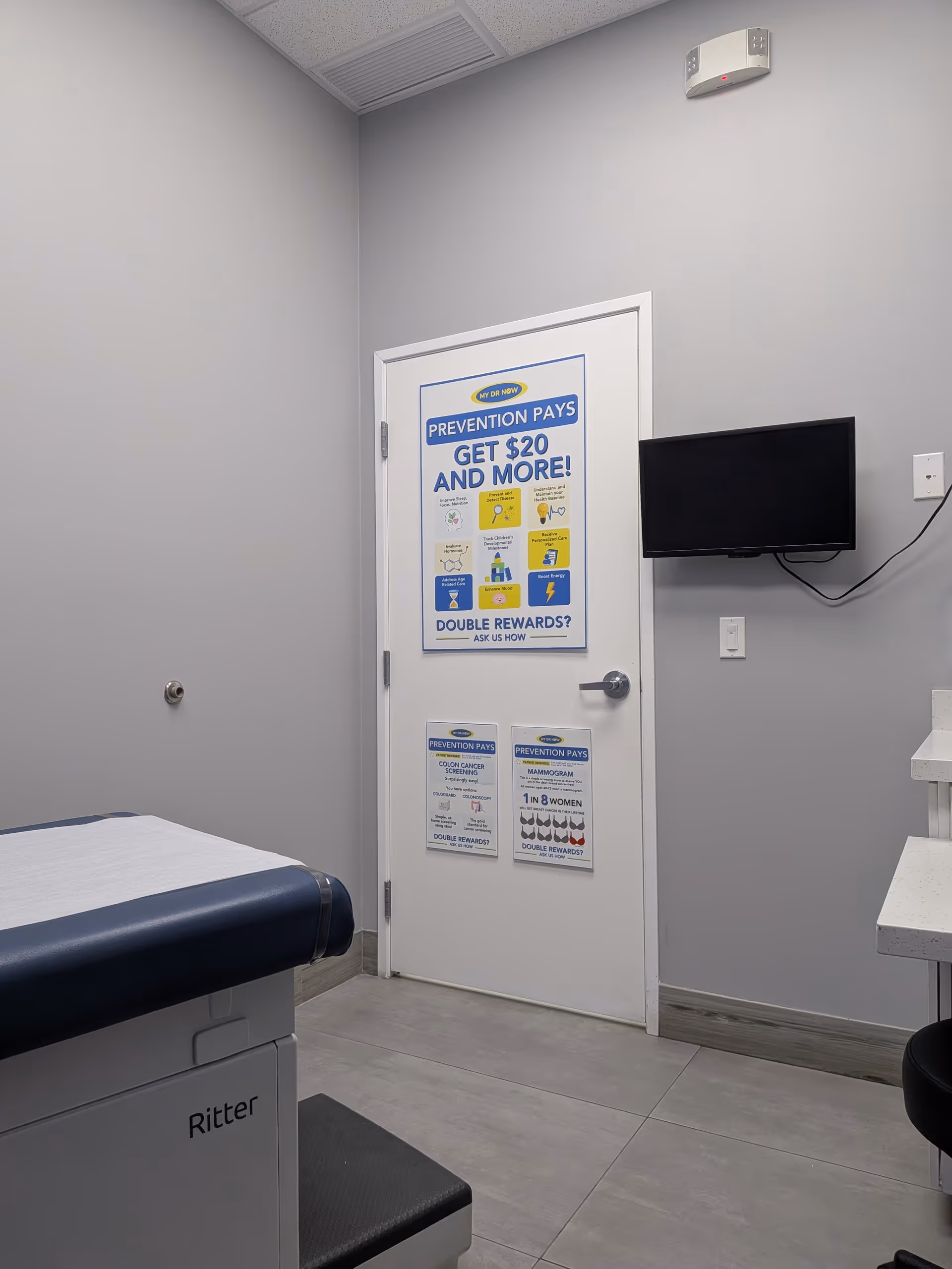 Medical exam room with a padded table, posters on a door, wall TV, and gray walls.