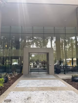 Front entrance of a modern glass building with a concrete walkway and landscaping.
