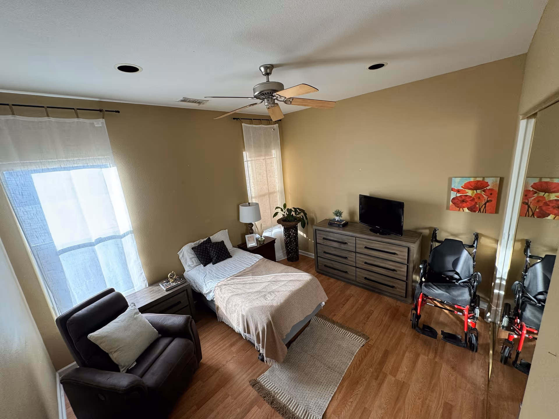 Senior living bedroom with bed, dresser, TV, couch, wheelchair, and warm beige walls.