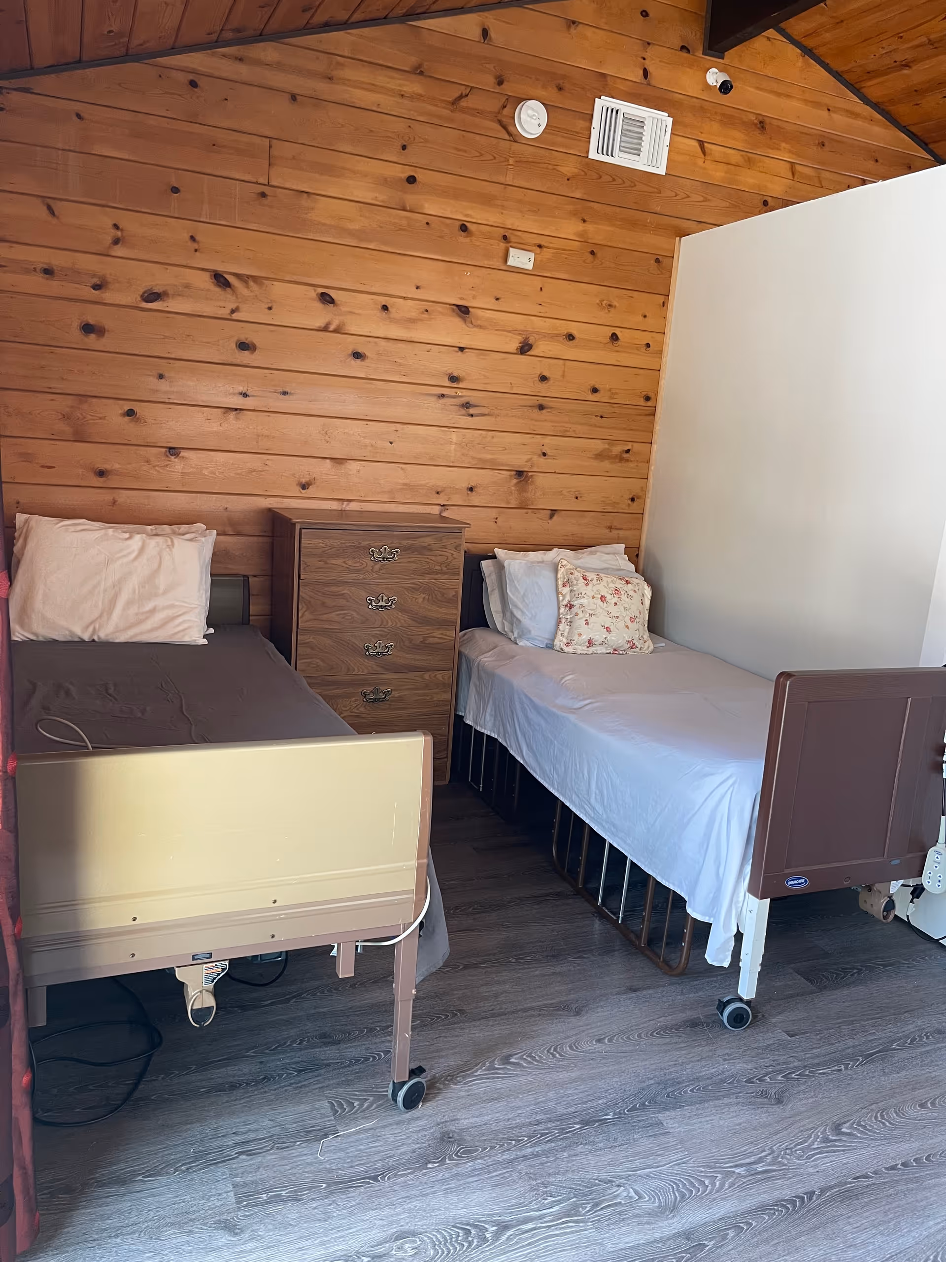 Two hospital-style beds with a wooden dresser in a wood-paneled resident bedroom.