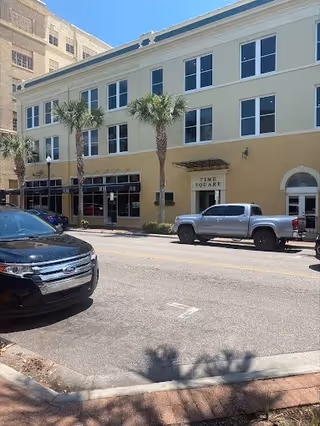 Exterior view of a multi-story building with storefronts, palm trees, and parked cars along the street.