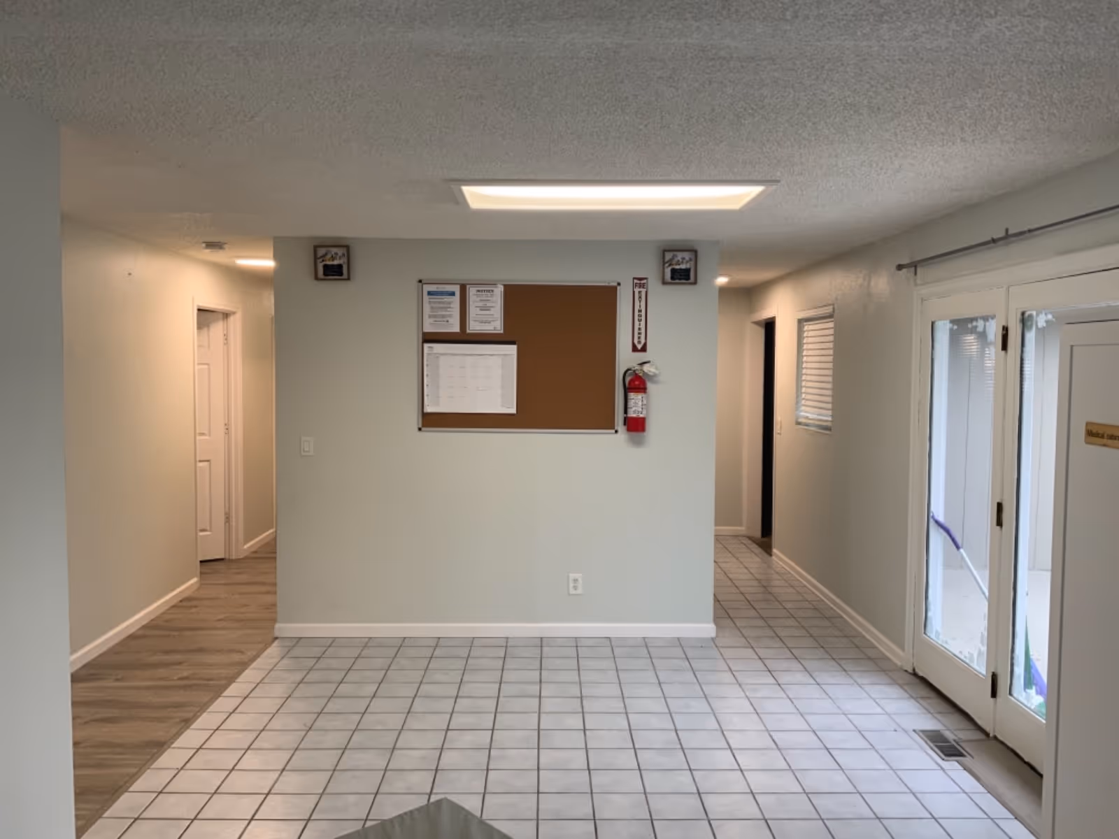 Bright, empty hallway with bulletin board, fire extinguisher, tiled floor, and glass doors.