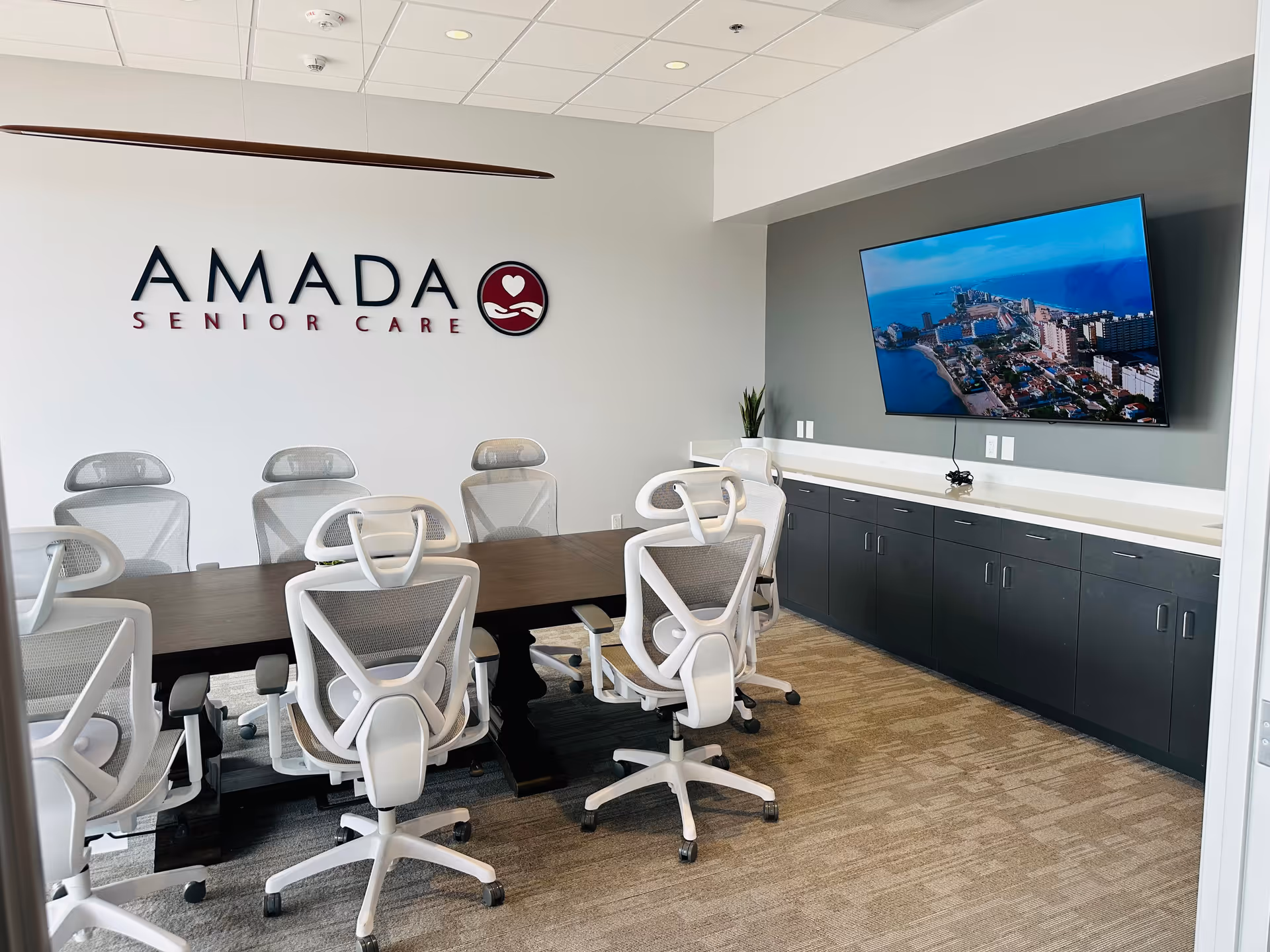 Modern conference room with white chairs, a long table, Amada Senior Care logo on the wall.