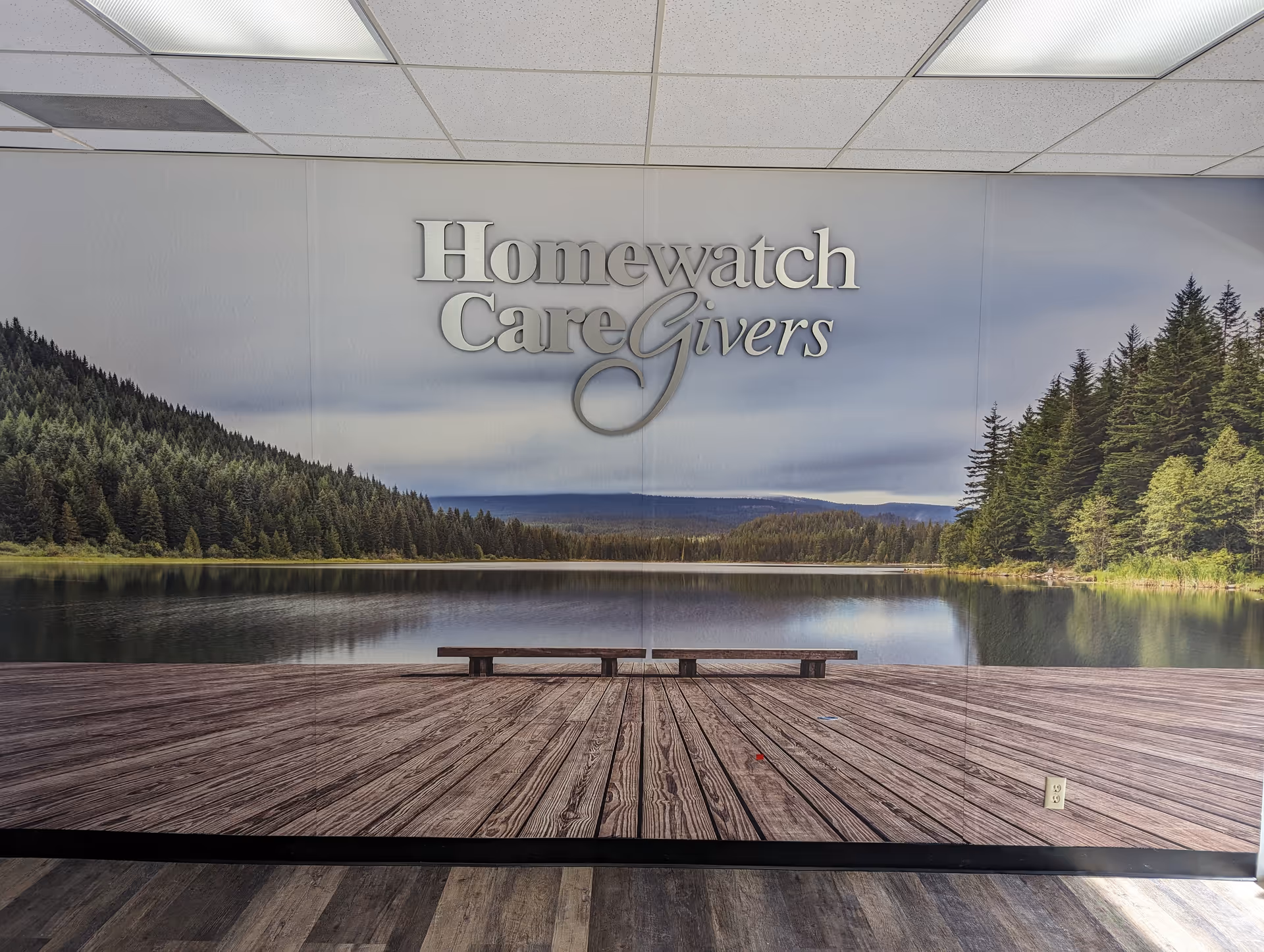 Indoor wall mural shows a lakeside scene with a wooden dock and the Homewatch CareGivers logo.