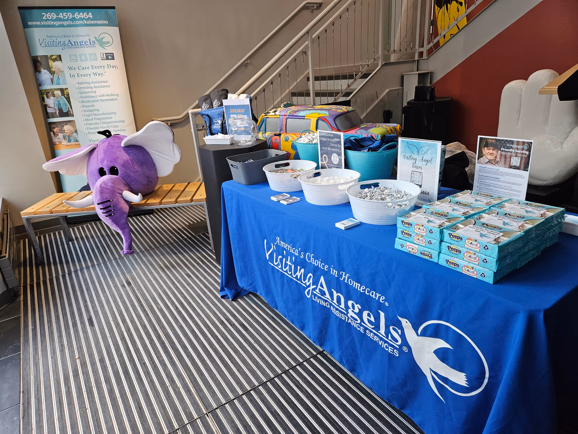 Indoor promotional booth with a blue Visiting Angels table, purple elephant mascot, and stairs.