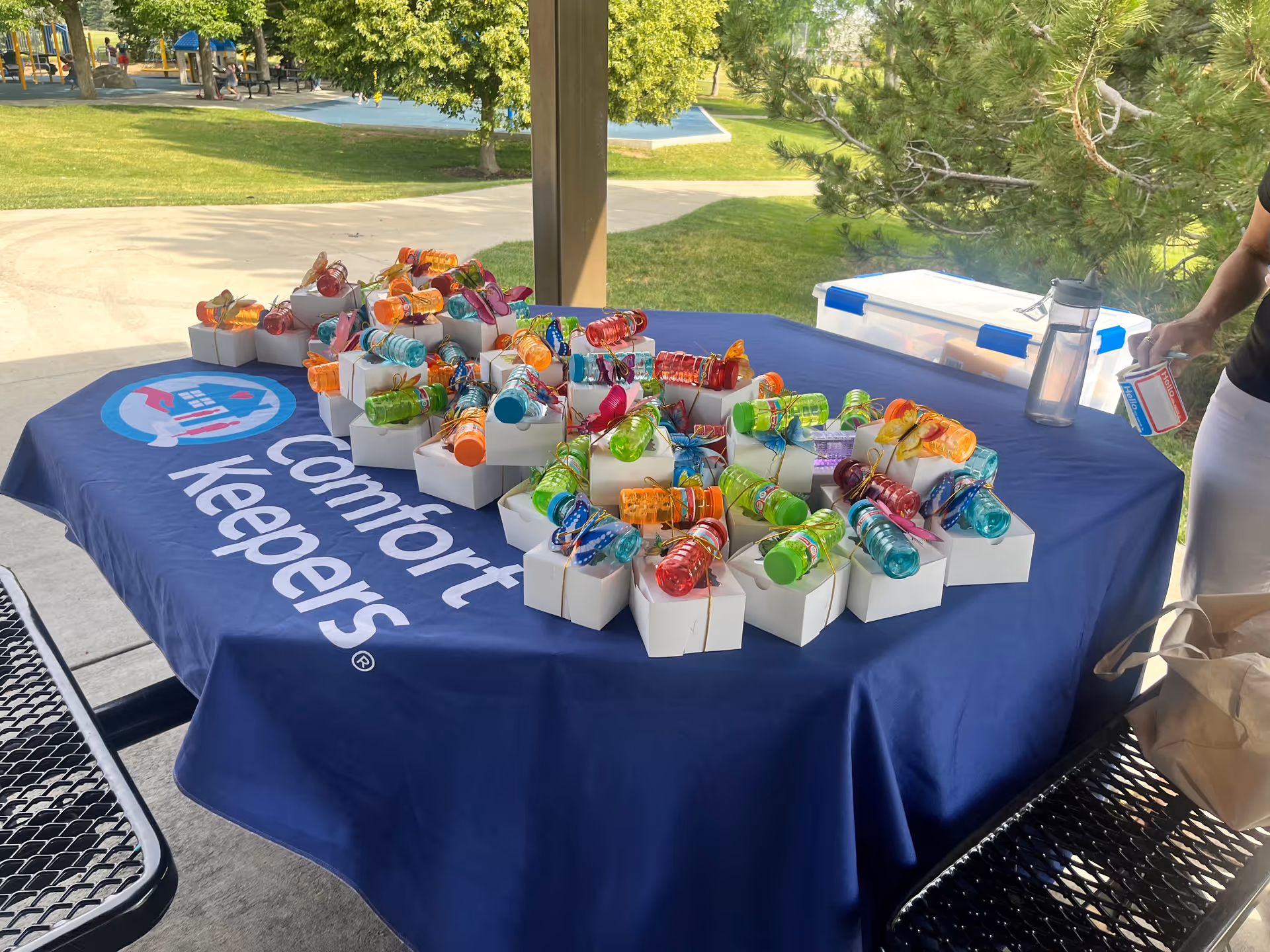 Outdoor tablecloth with blue Comfort Keepers branding and colorful gift boxes under a pavilion.
