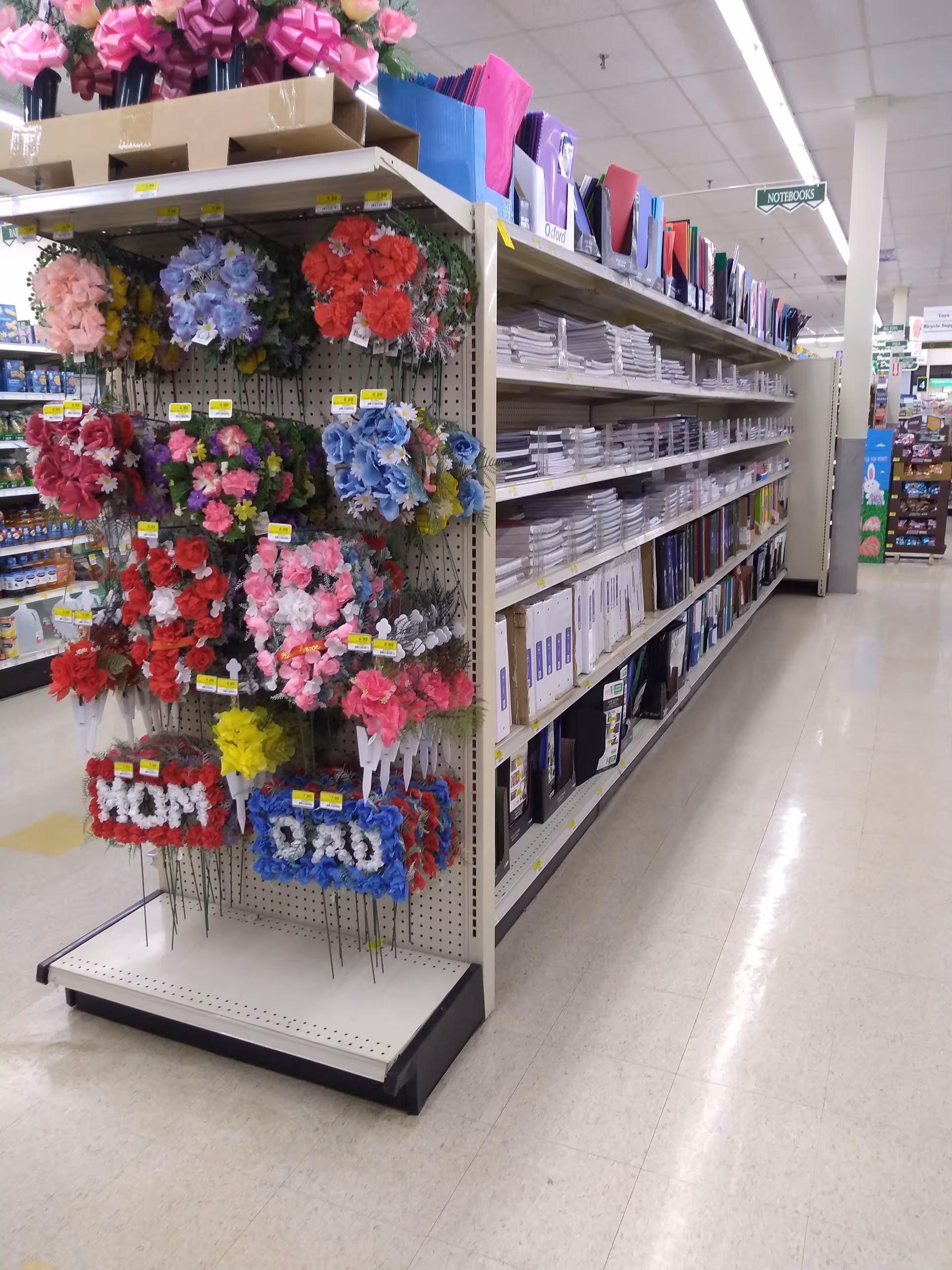 Aisle in Discount Drug Mart store with shelves of notebooks and colorful artificial flowers.
