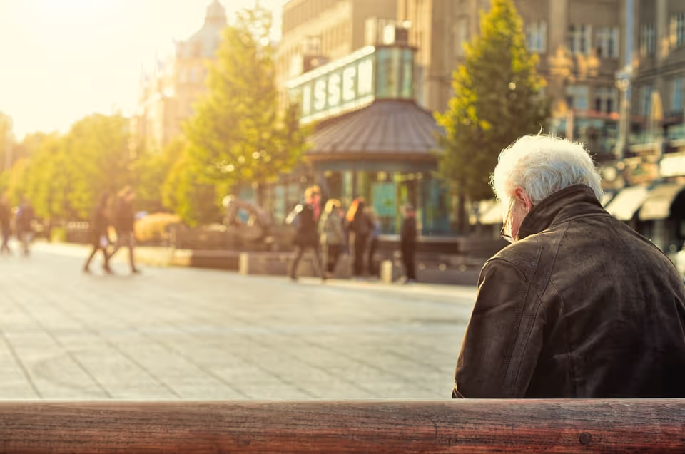 An elderly man sits on a wooden bench in a sunny urban plaza.