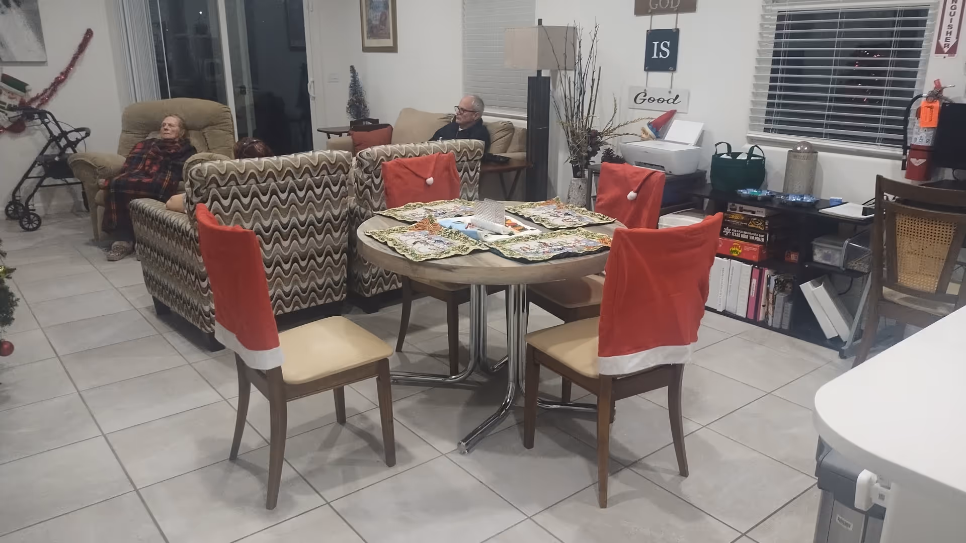 Senior living common room with round dining table, red chair covers, and residents relaxing.