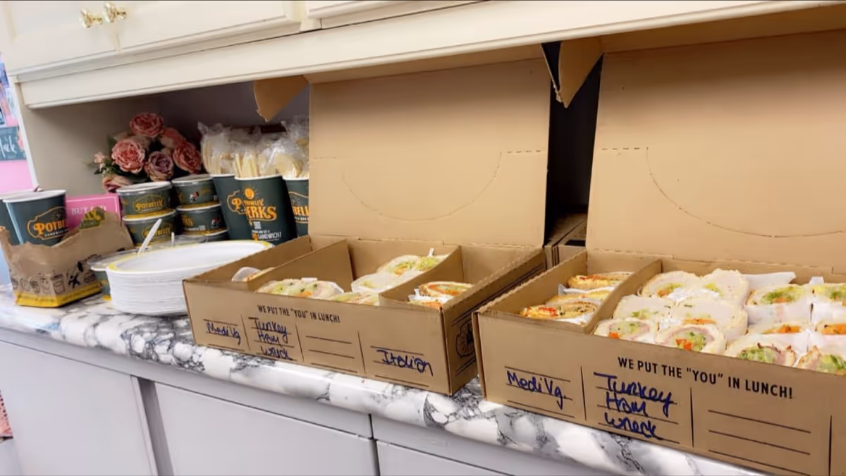 Boxes of sandwiches on a marble counter with cups and flowers in a kitchen.