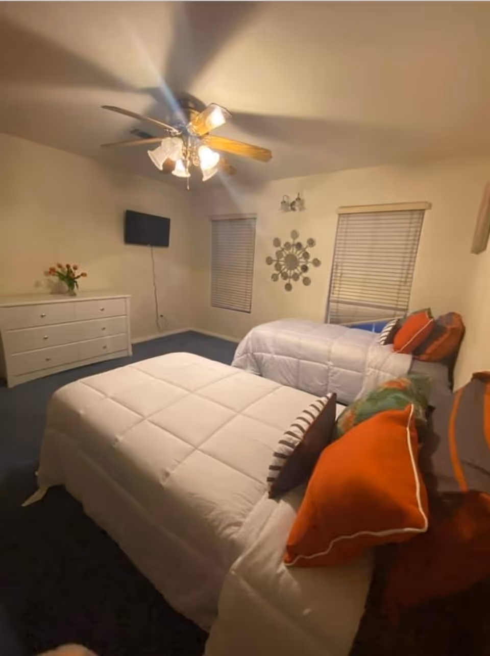 Two neatly made beds in a light-colored bedroom with dresser and ceiling fan.