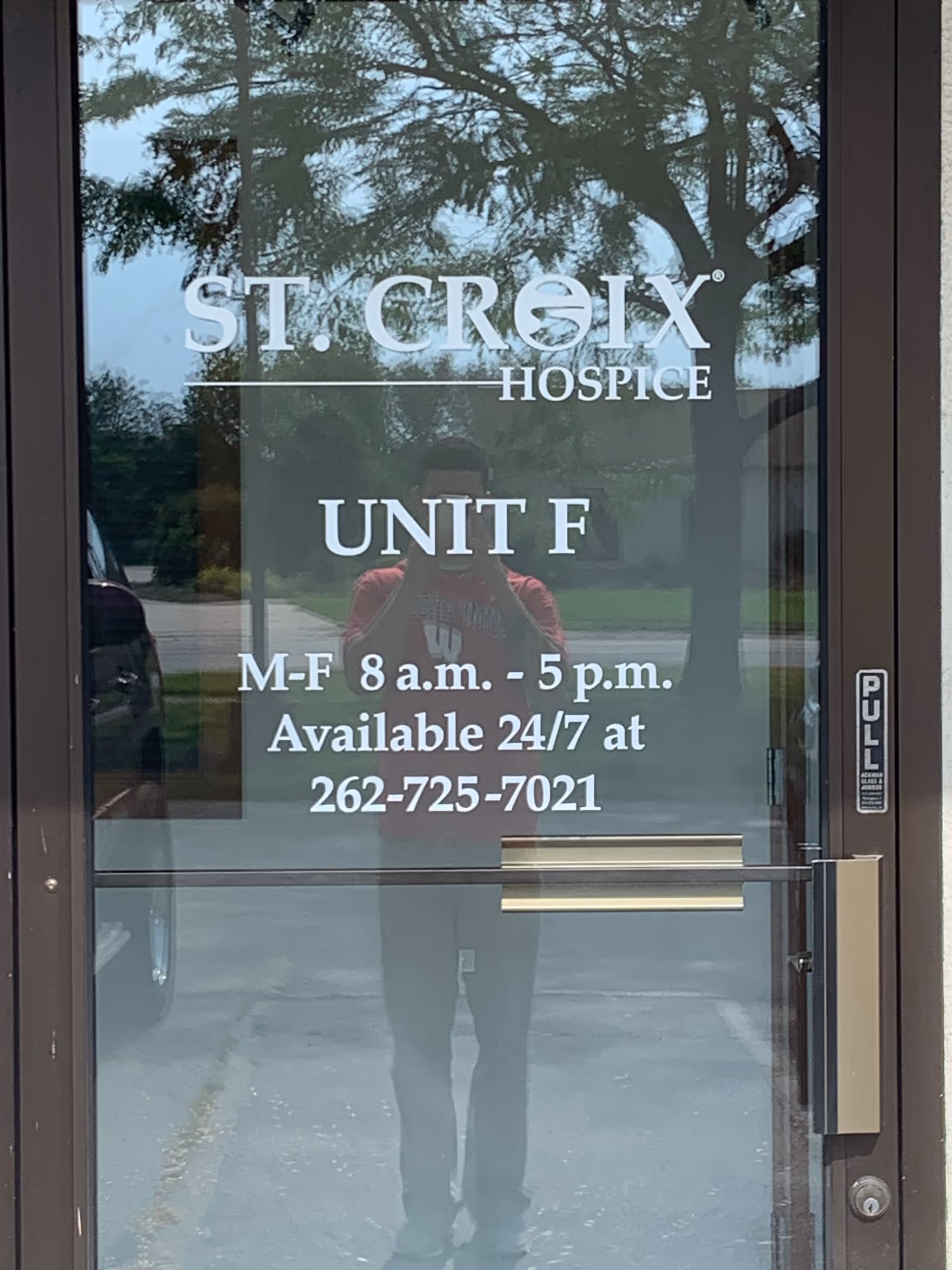 Glass entrance door shows St. Croix Hospice Unit F signage hours; reflection of trees and person.