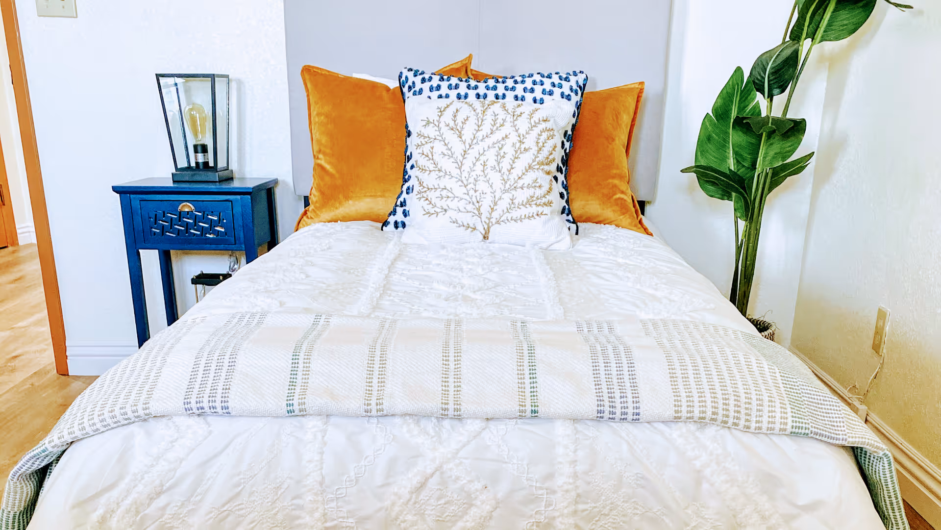 A neatly made bedroom with a bed, blue nightstand, orange pillows, and a tall plant.