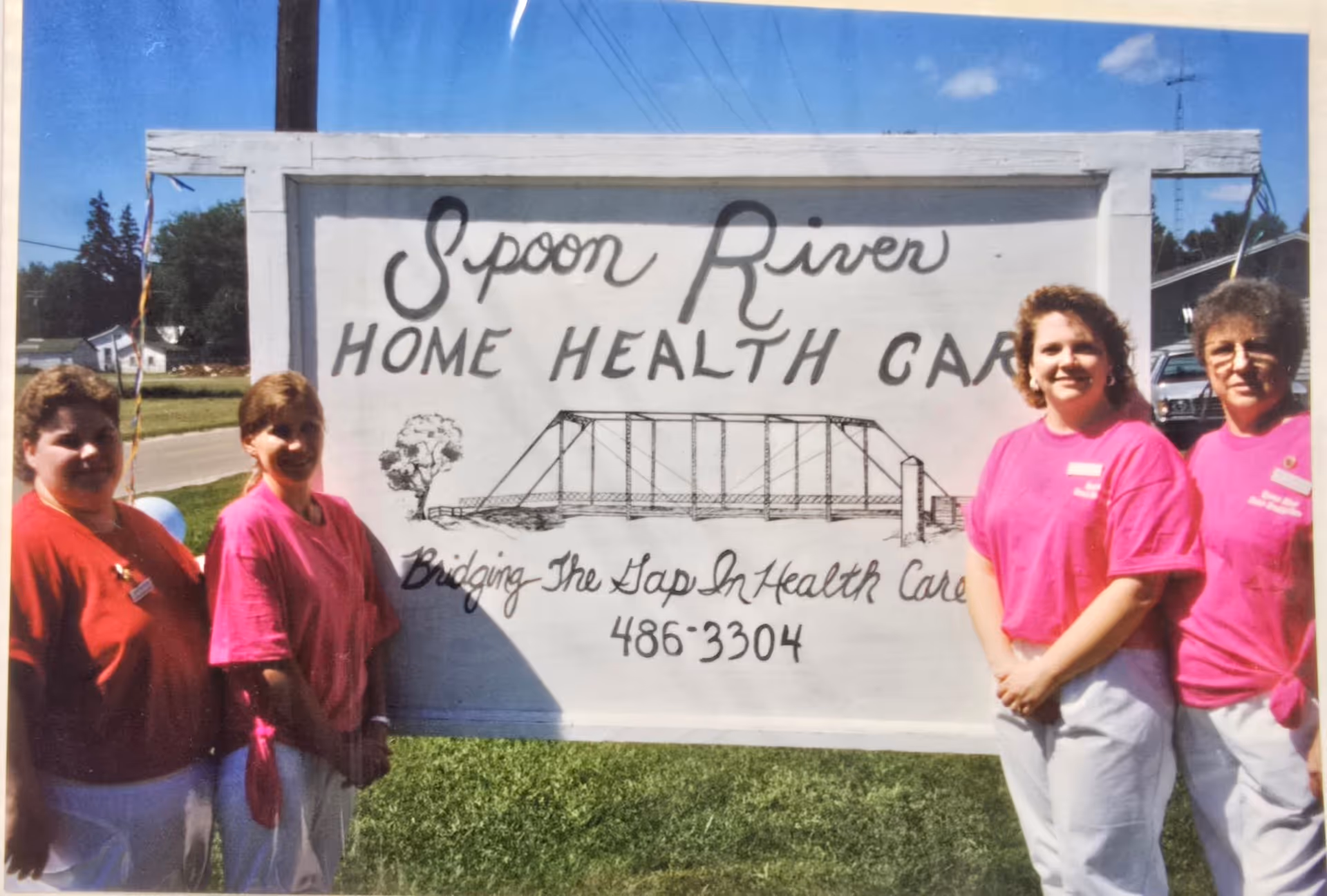 Four staff members stand outdoors beside a Spoon River Home Health Care sign with bridge illustration.
