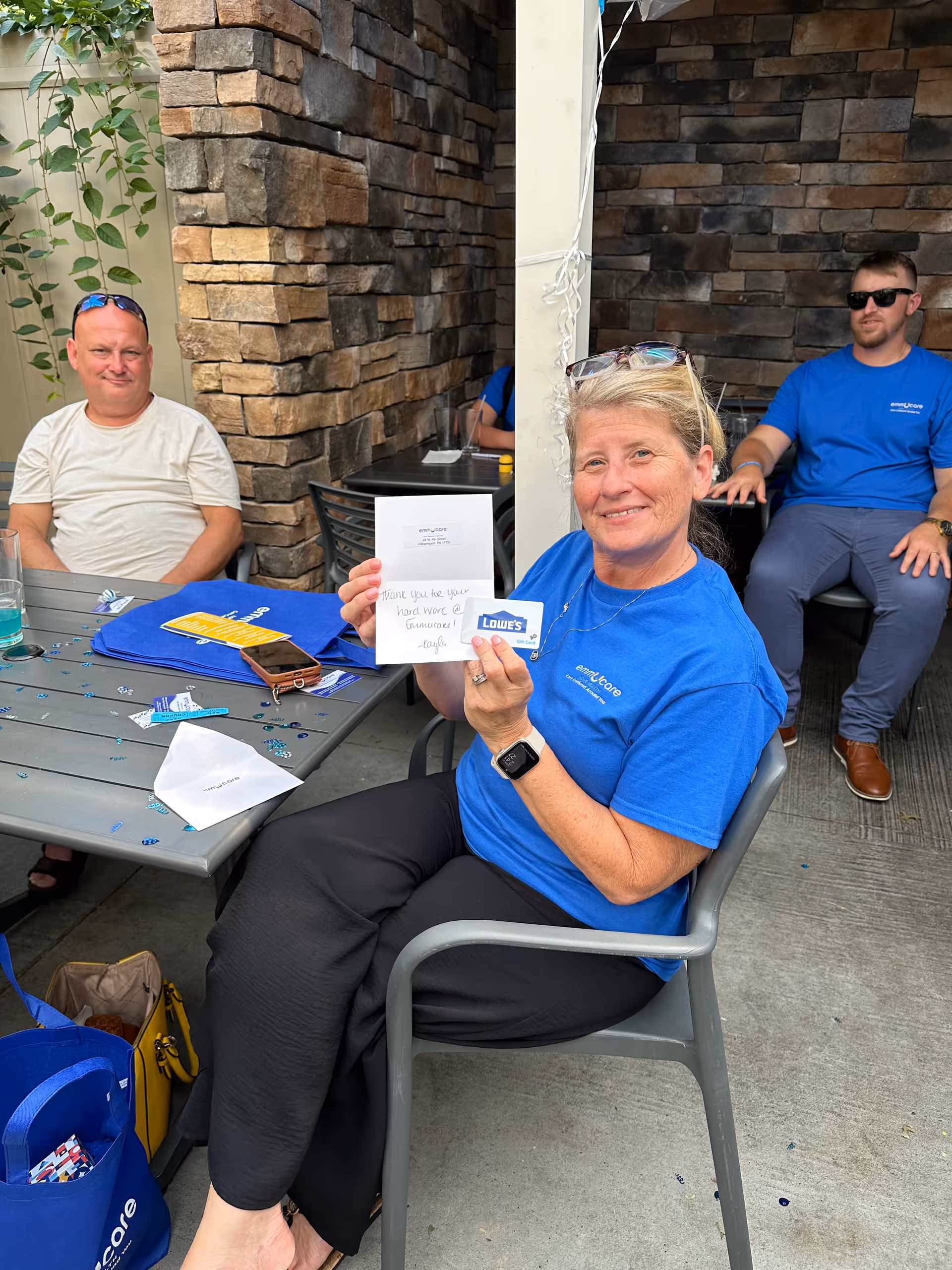 Two caregivers in blue shirts sit outdoors with documents at a stone-walled patio.