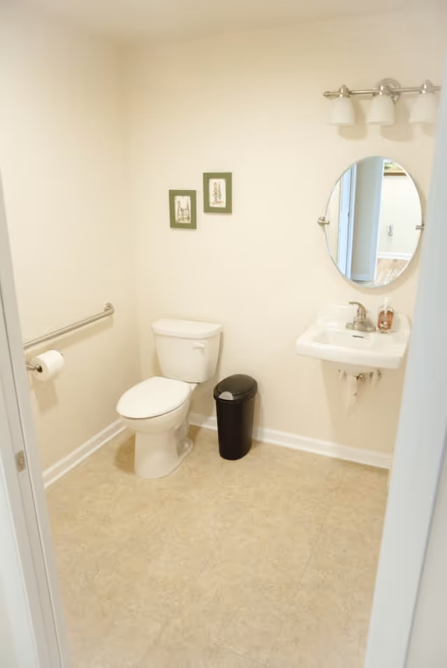 Small white bathroom with toilet, wall-mounted sink, oval mirror, and grab bar.