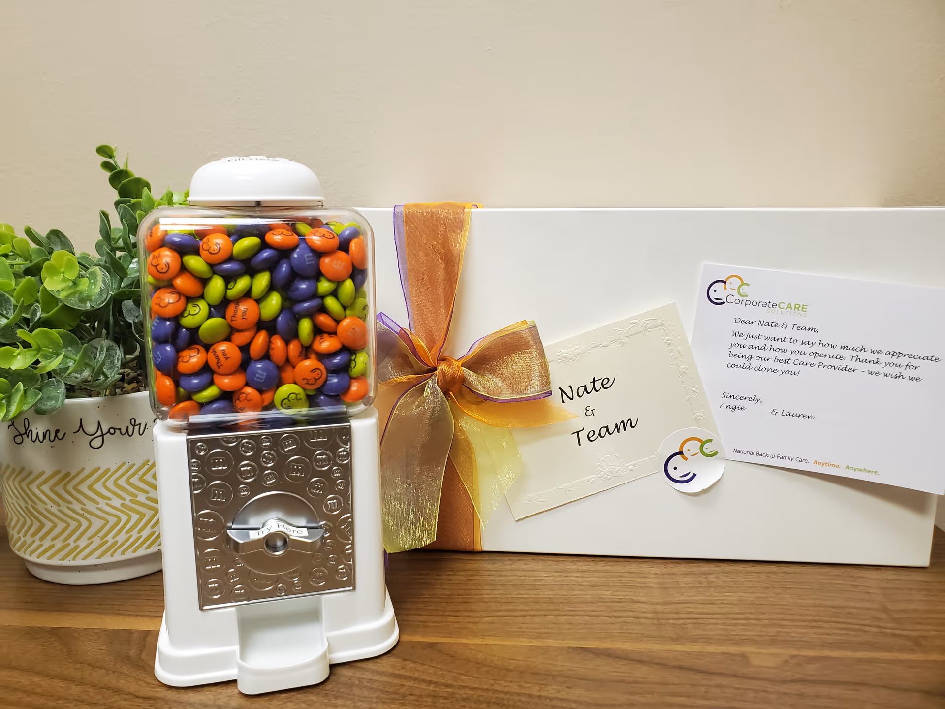 Candy dispenser filled with colorful gumballs beside a wrapped gift on a wooden counter.