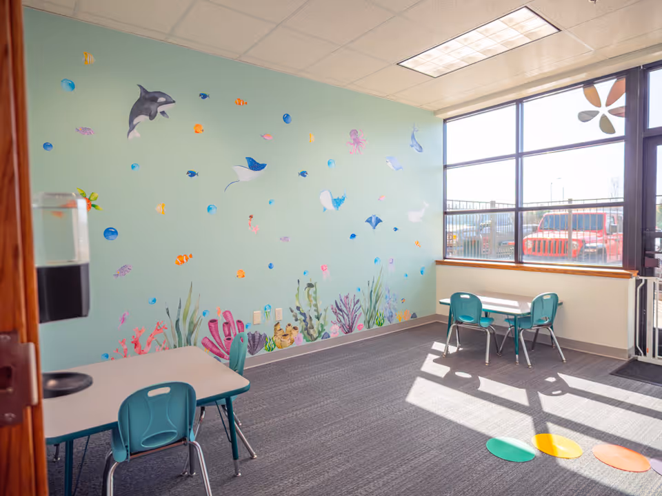 Colorful interior playroom with aquatic mural, small tables, chairs, and large window.