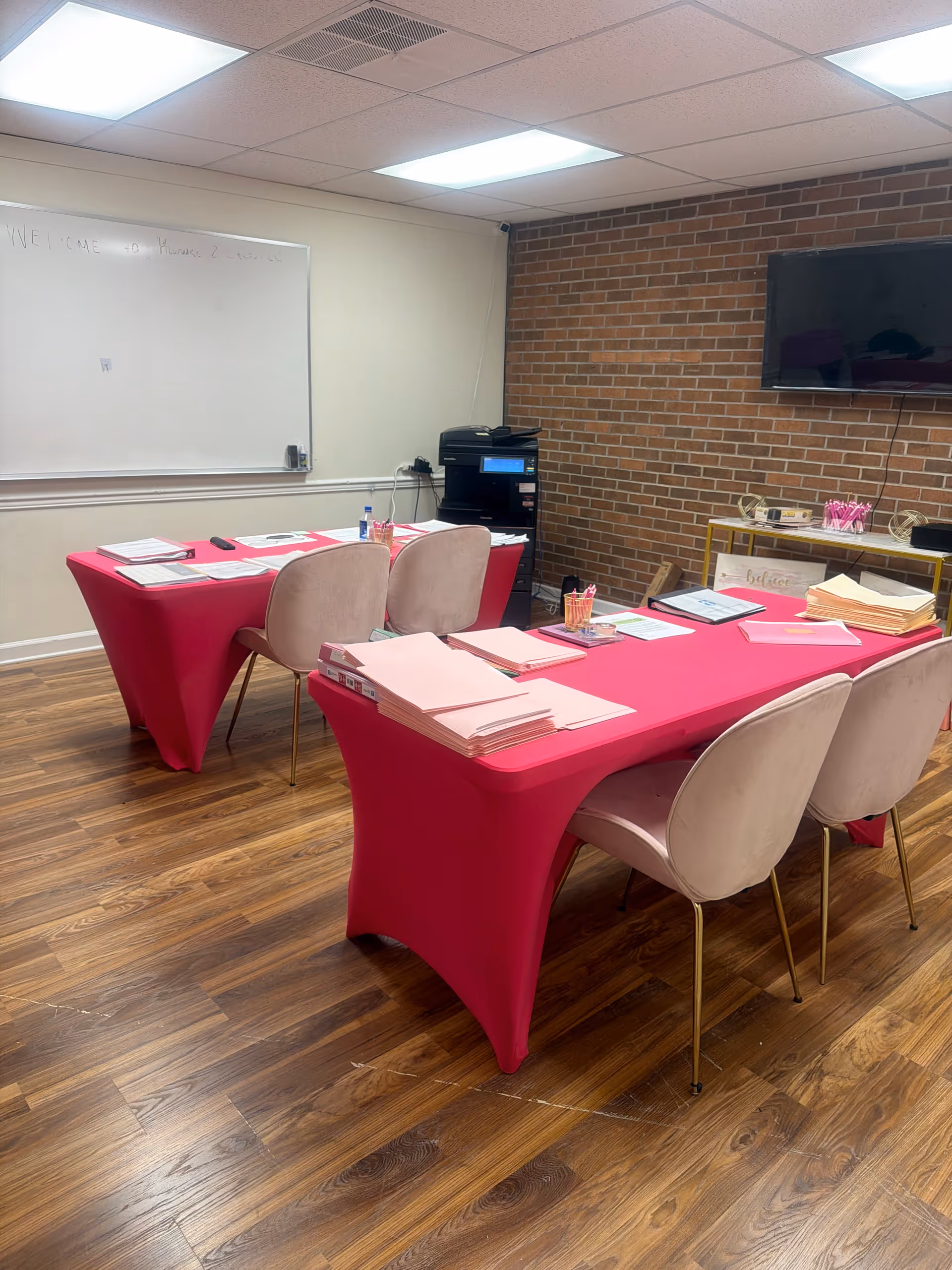 Interior meeting room with pink tablecloths, stacks of papers, and beige chairs.