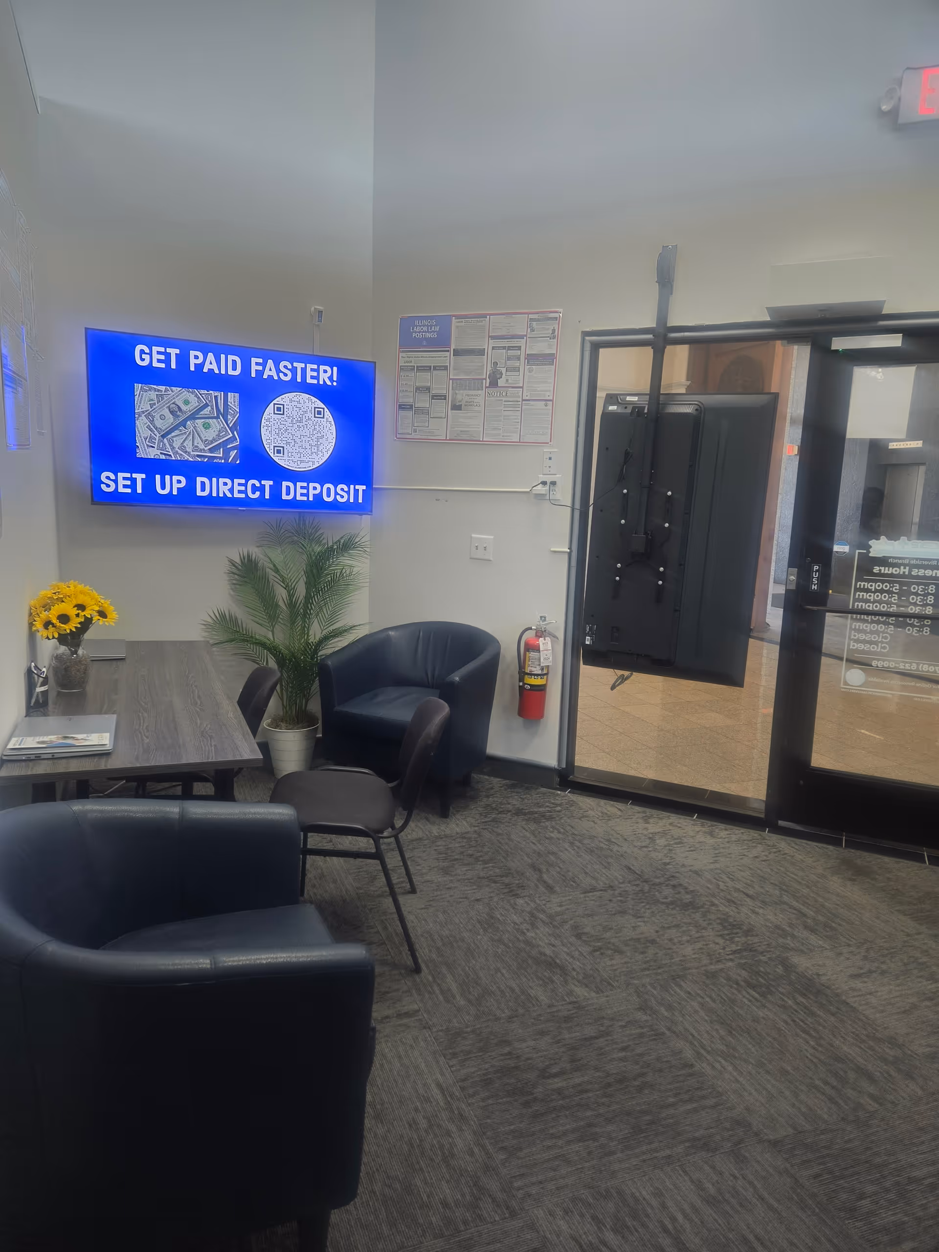 Interior waiting area with a blue digital sign, seating, plant, and glass doors.