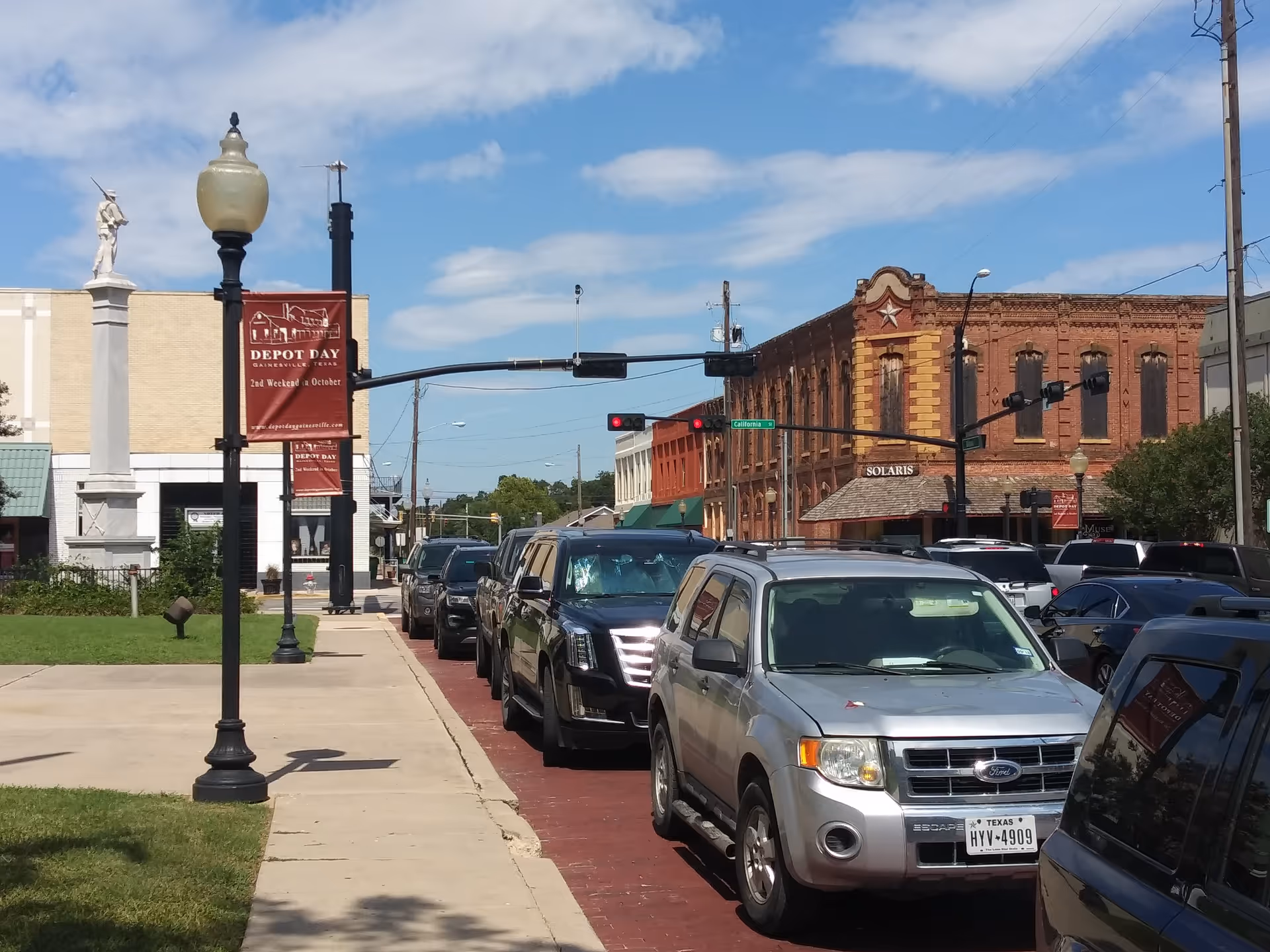 Outdoor street scene with parked cars and brick storefronts under blue sky.