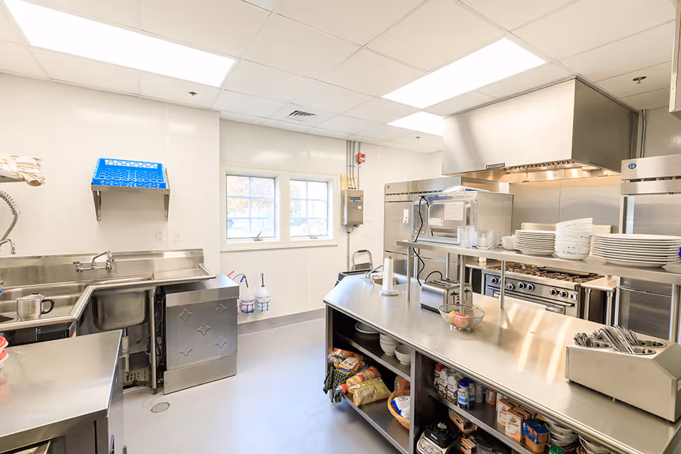 Commercial-style kitchen in a senior living facility with stainless steel counters, ovens, and dishware.