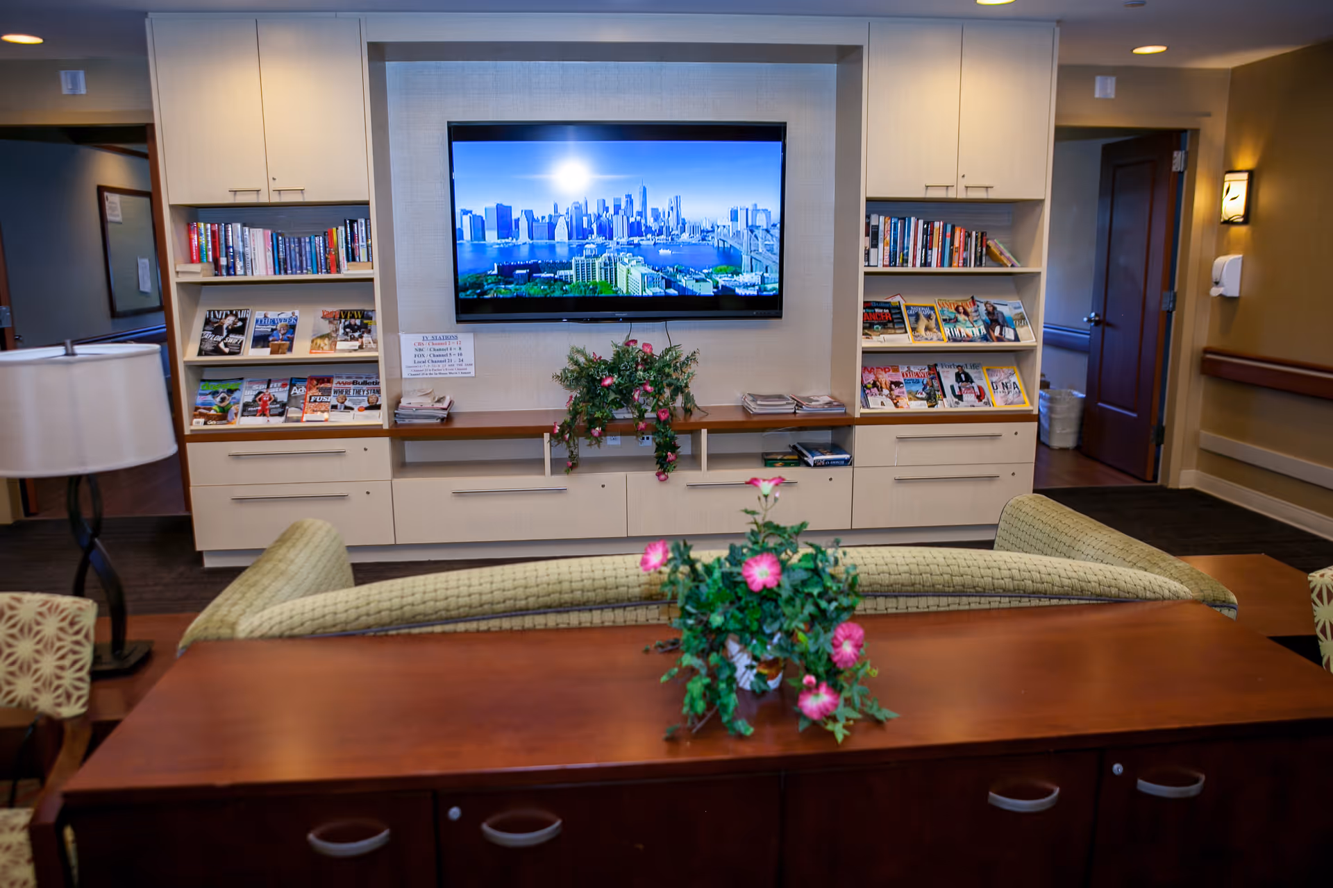 Senior living facility lounge featuring a wall TV, bookshelves, and floral arrangements