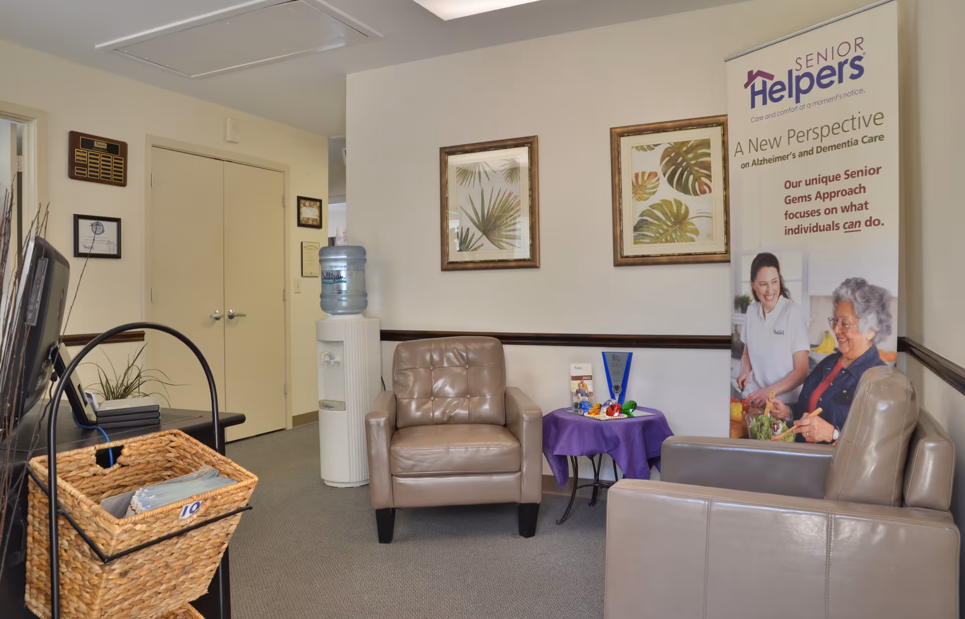 Waiting area with beige chairs, a water cooler, small table, and Senior Helpers banner.