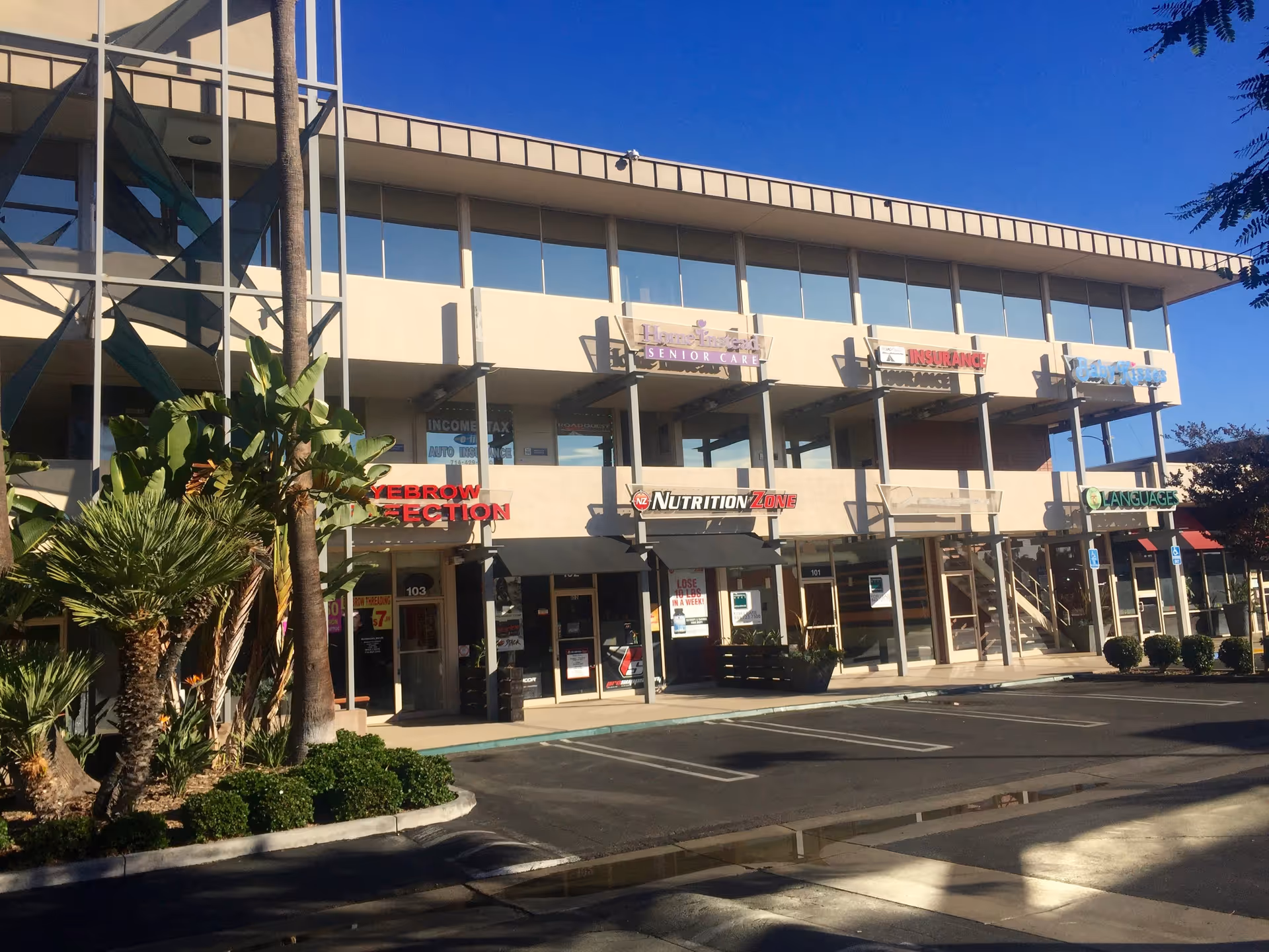 Two-story strip mall exterior with glass balconies, palm trees, and various storefronts.