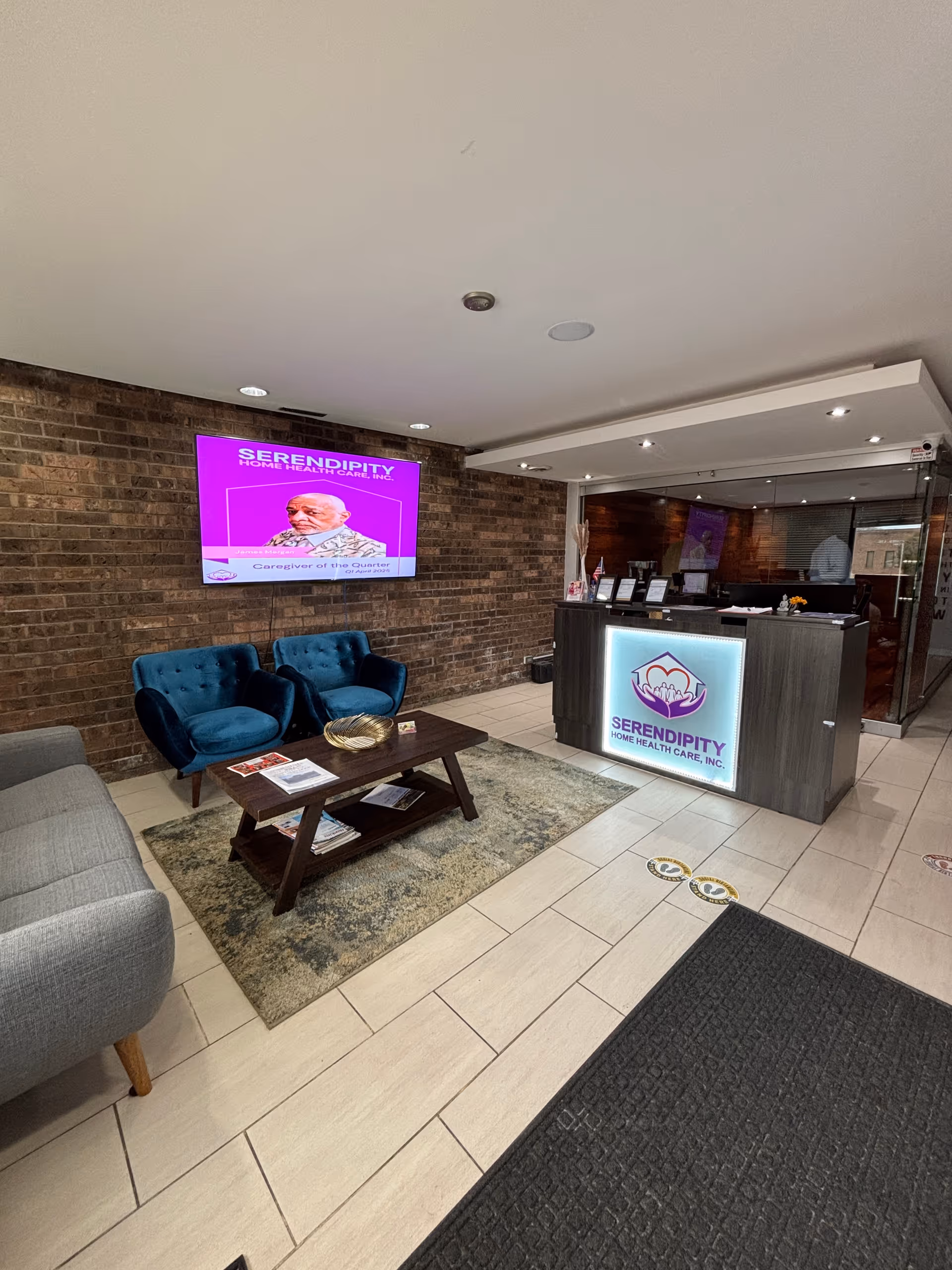 Lobby area with brick wall, mounted screen, reception desk, and blue upholstered chairs.