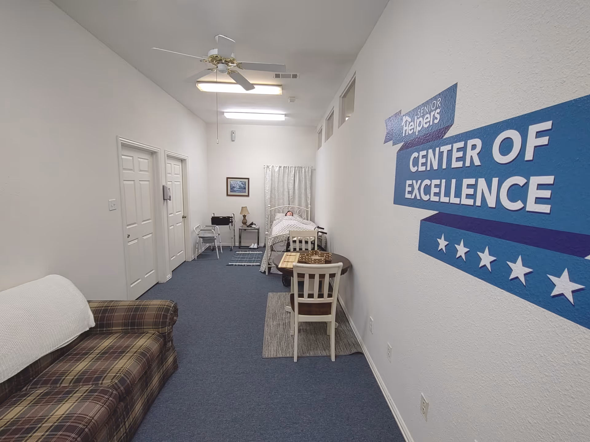 Inside Senior Helpers facility hallway, featuring bed, seating, and a large Center of Excellence sign.