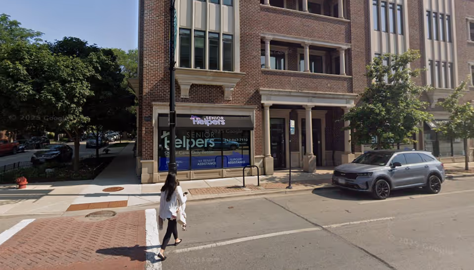 Brick building with Senior Helpers storefront on a city street; pedestrian nearby and a parked SUV.