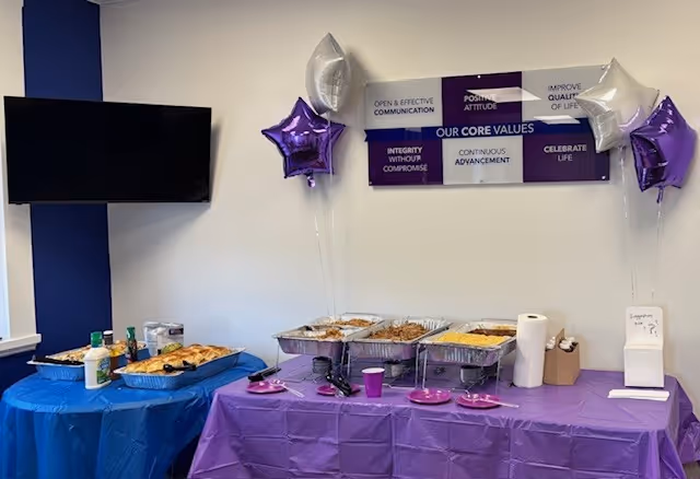 Buffet table with chafing dishes, purple and blue cloths, balloons, inside a dining room.