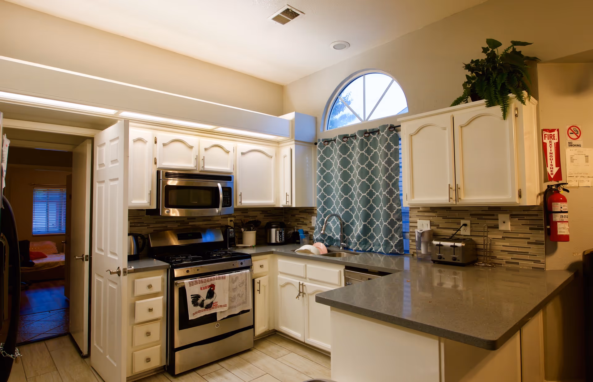 White kitchen with stainless appliances, grey countertops, and teal patterned curtain over the sink.