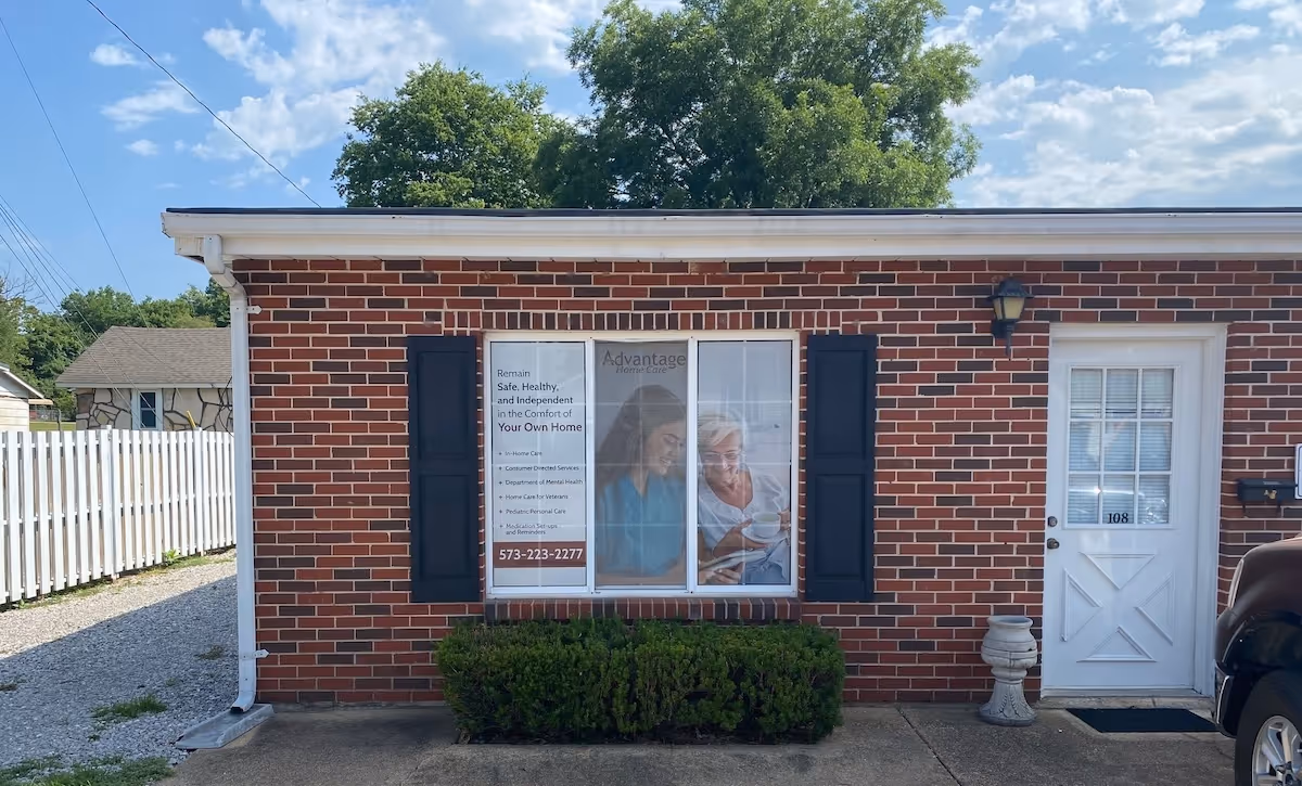 Brick building front with a window display, black shutters, white door, and shrub in front.