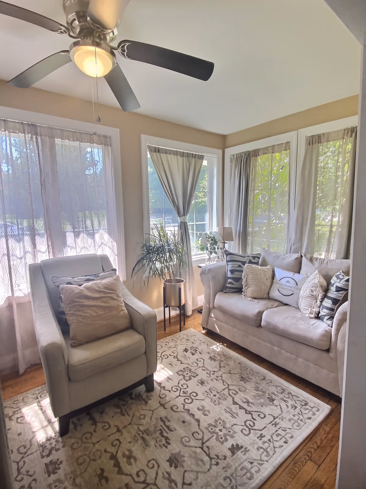 Bright living room with beige furniture, plants, and large windows with sheer curtains.