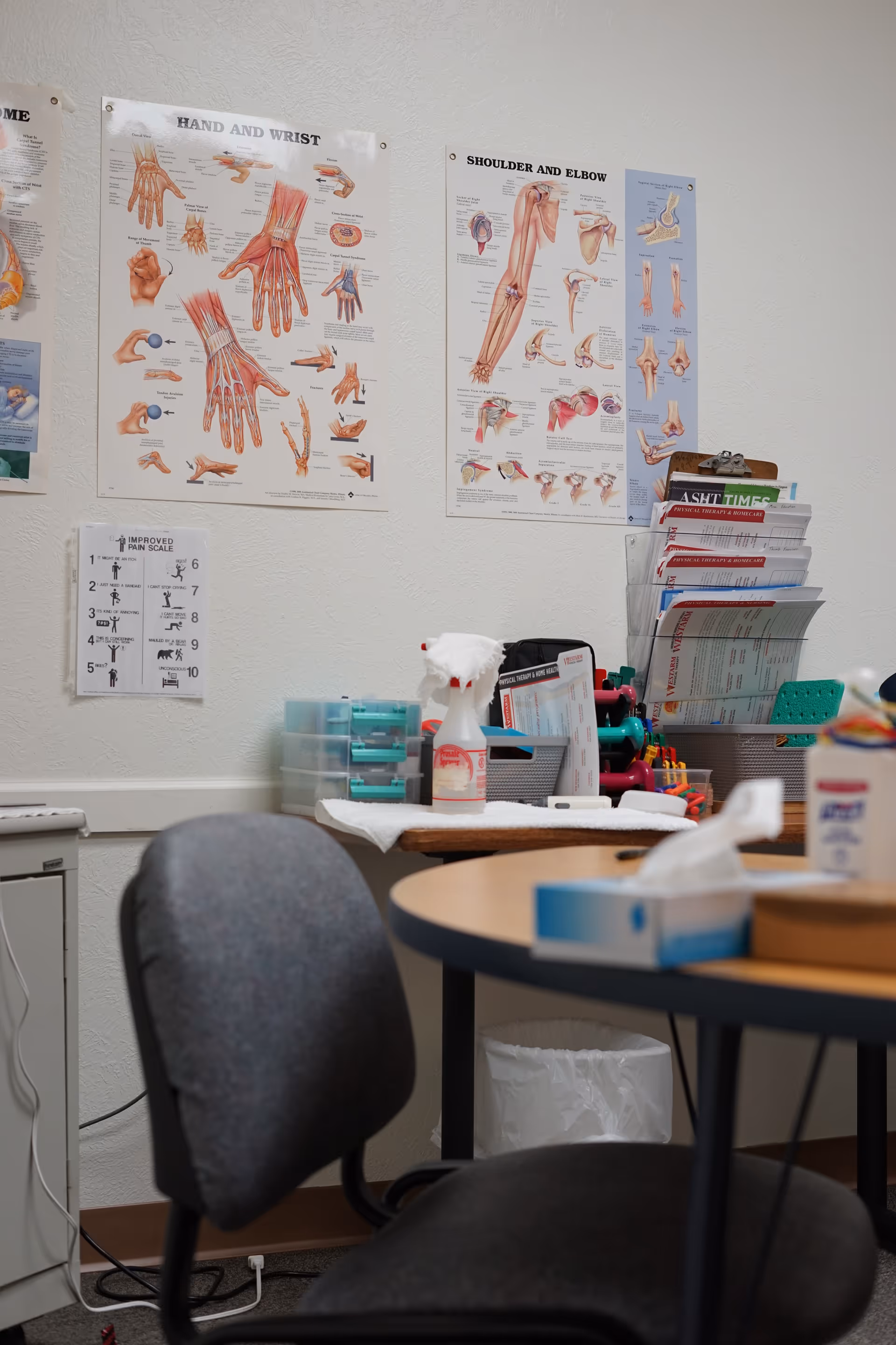Interior clinic room with anatomy posters, a round table, chair, and supplies.
